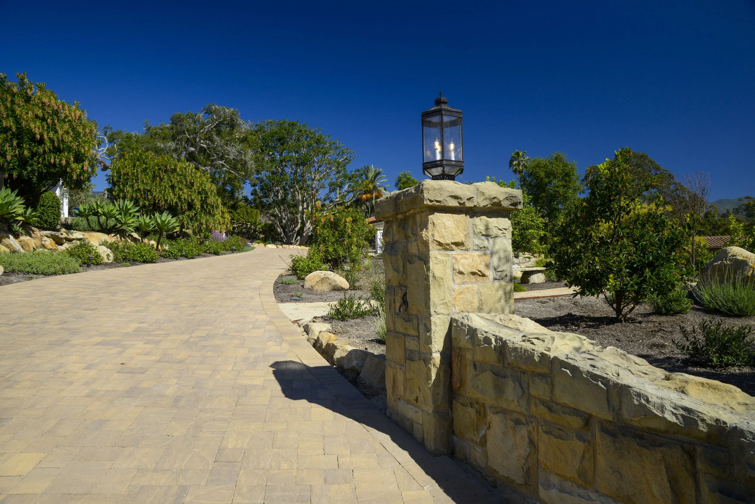 A paved sidewalk in a garden with green bushes, trees, and a stone wall with a lamp post on top under a clear blue sky.