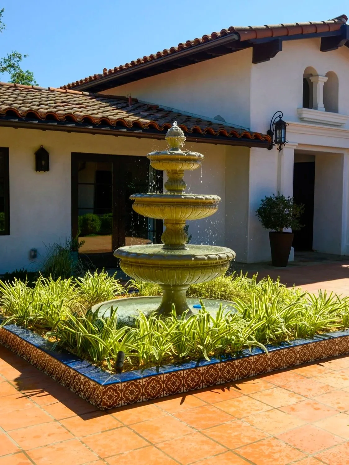 A stone fountain with three tiers surrounded by green plants in a decorative tile border in front of a white house with red-tiled roof.