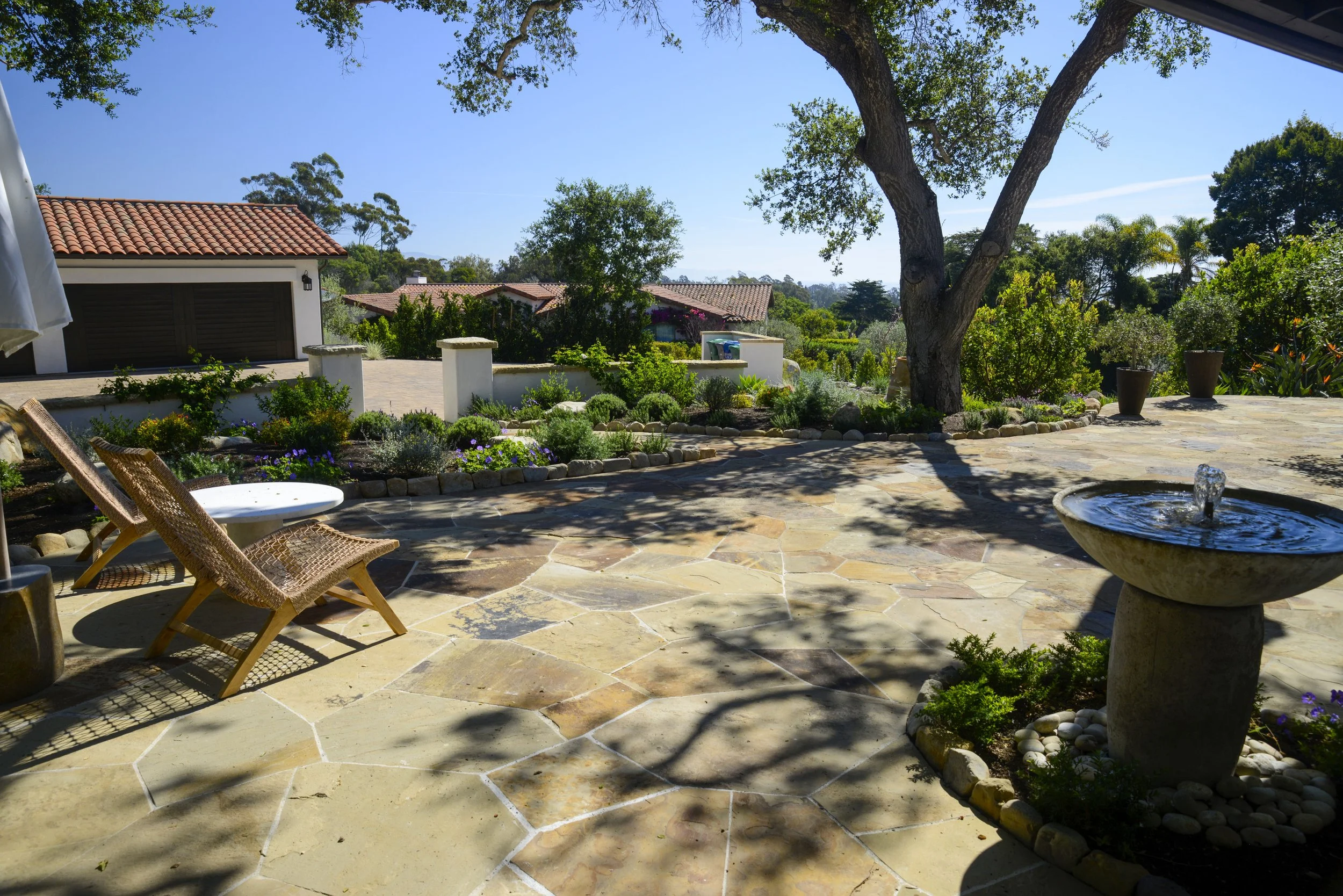 Sunny backyard patio with stone flooring, a large tree casting shadows, potted plants, a water fountain, and a view of neighboring houses with tiled roofs under a clear blue sky.