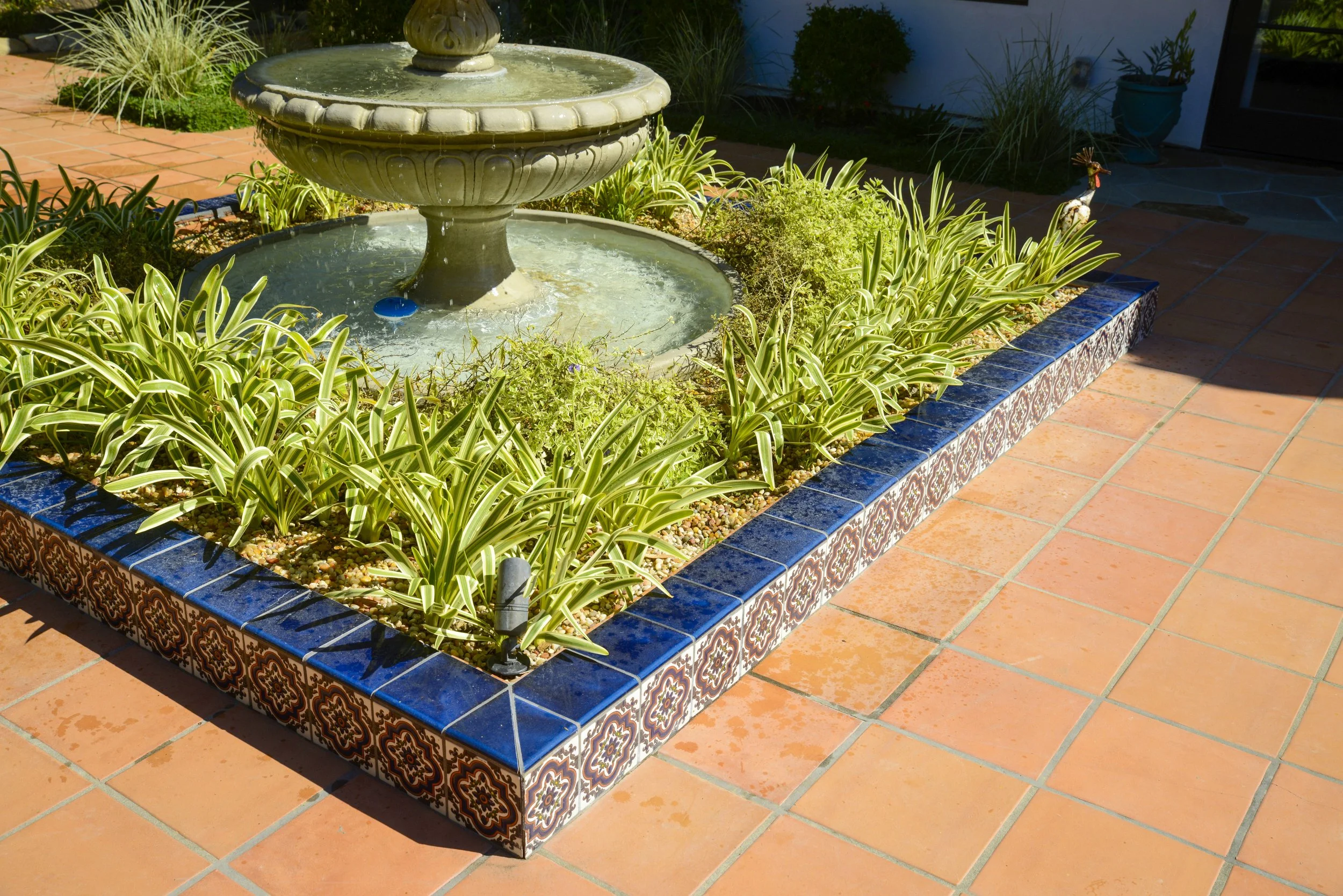 A decorative garden with a small fountain at the center, surrounded by green and yellow plants, bordered with blue and patterned tiles, and set on terracotta tile flooring.