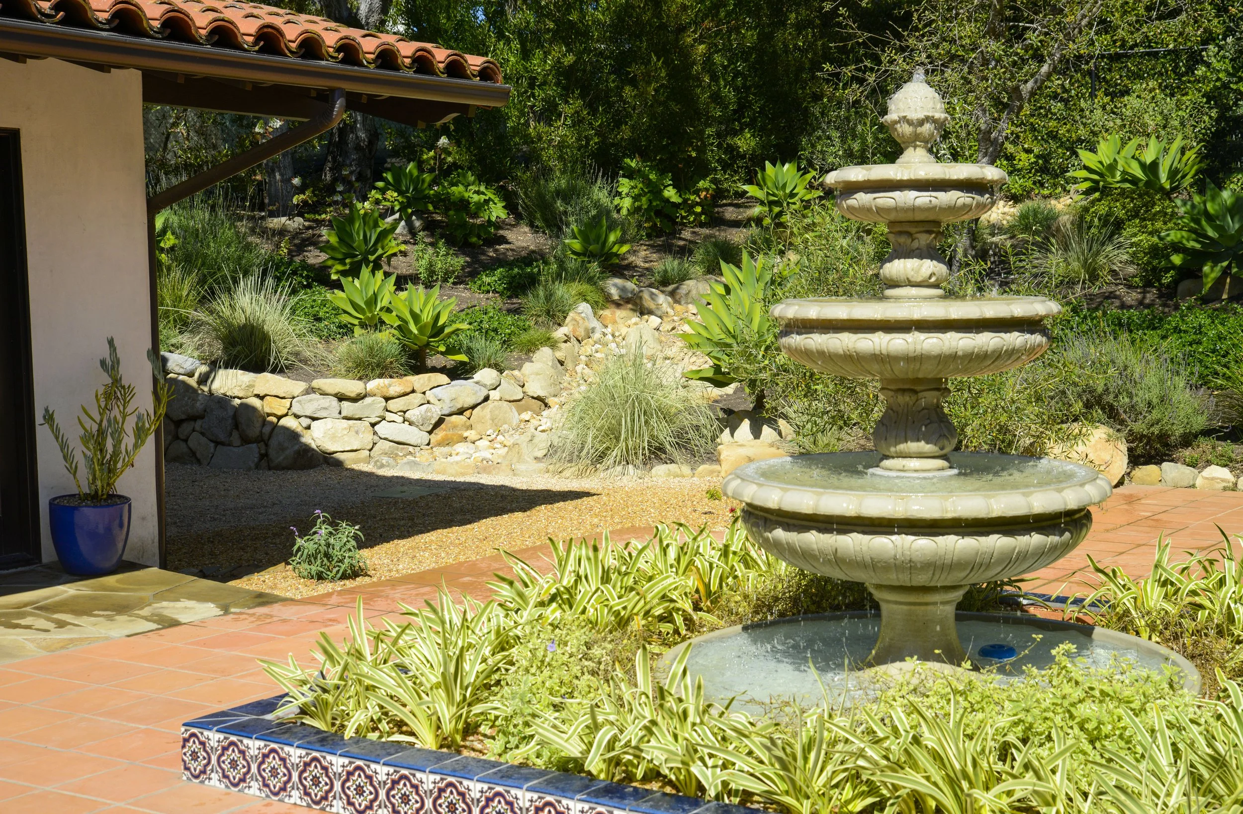 A multi-tiered stone fountain in a garden with green plants, succulents, and a stone border, next to a patio with a potted plant in a blue container.
