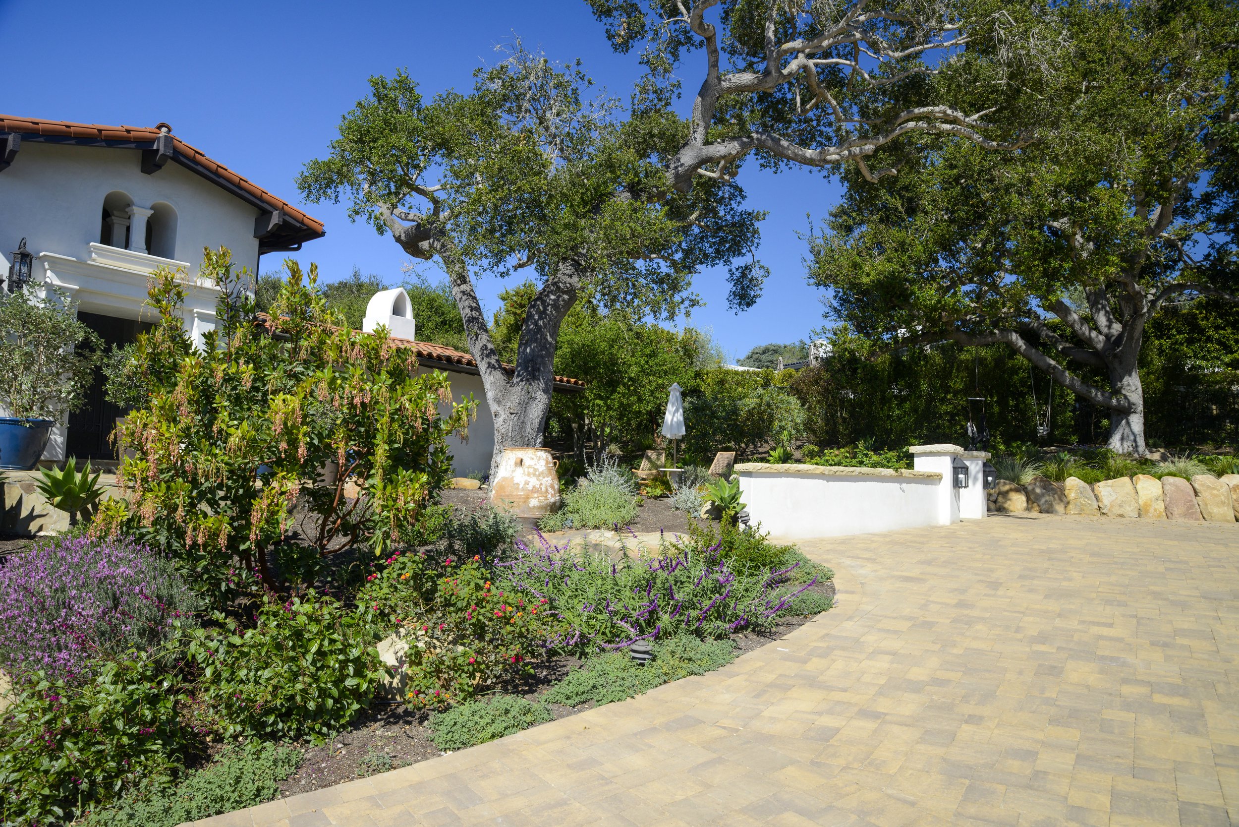 A paved driveway leading to a garden with flowers and large trees, adjacent to a white house with a tile roof and a chimney, under a clear blue sky.