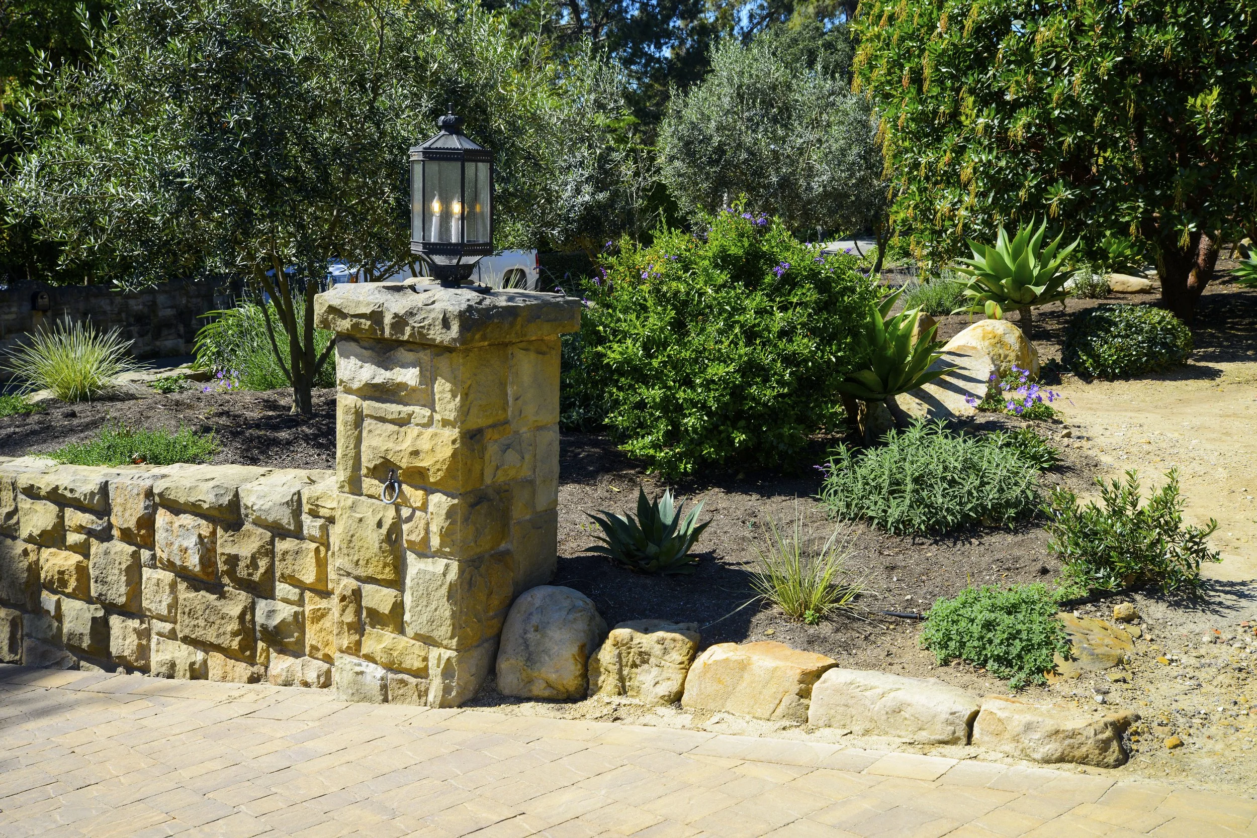 A landscaped garden area with a stone wall and pillar, topped with a black lantern, surrounded by various green plants and succulents, with a tree in the background and a dirt pathway.