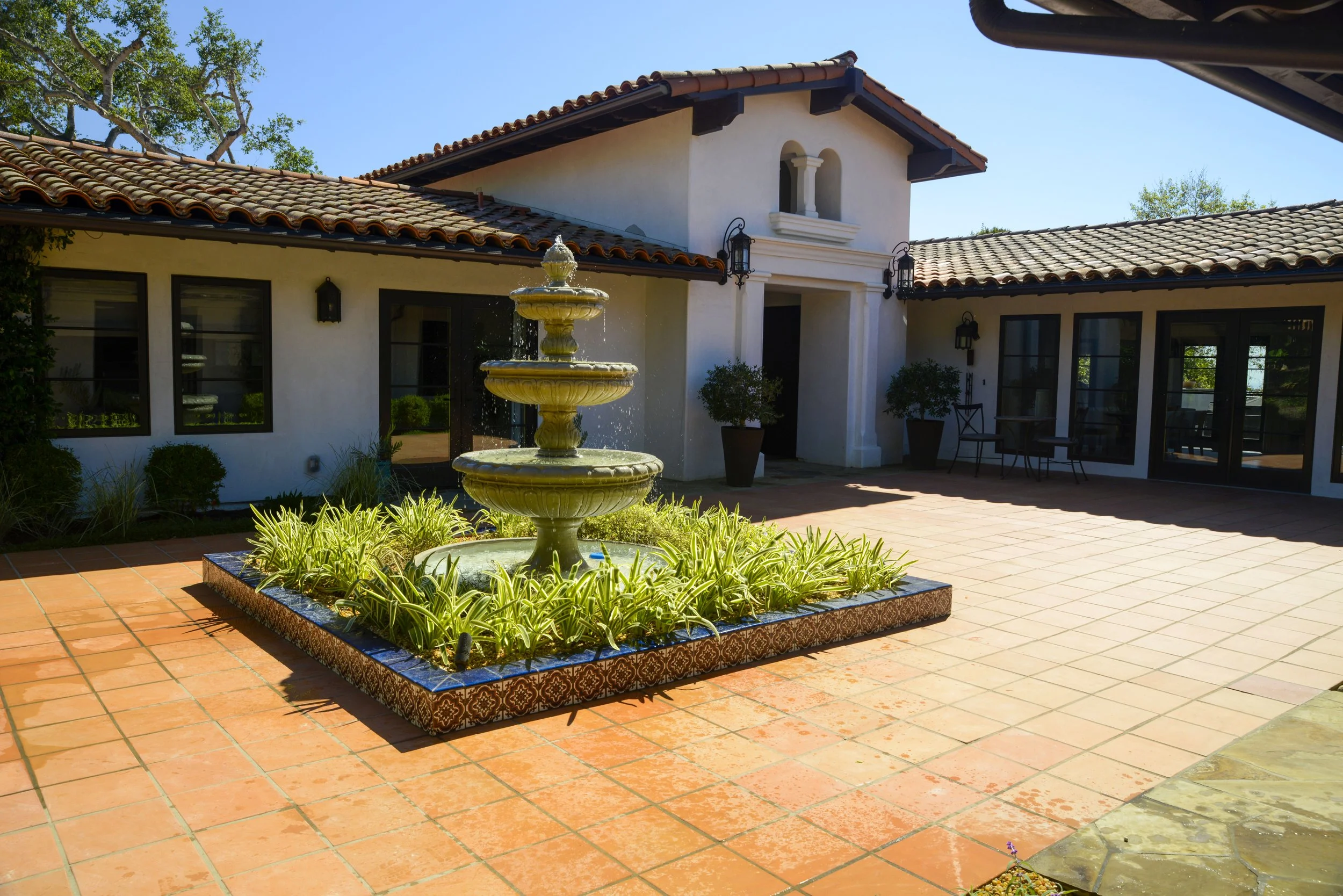 A courtyard with a three-tiered fountain surrounded by plants, terracotta tile flooring, two white stucco buildings with Spanish-style red tile roofs, and patio furniture, under a clear blue sky.