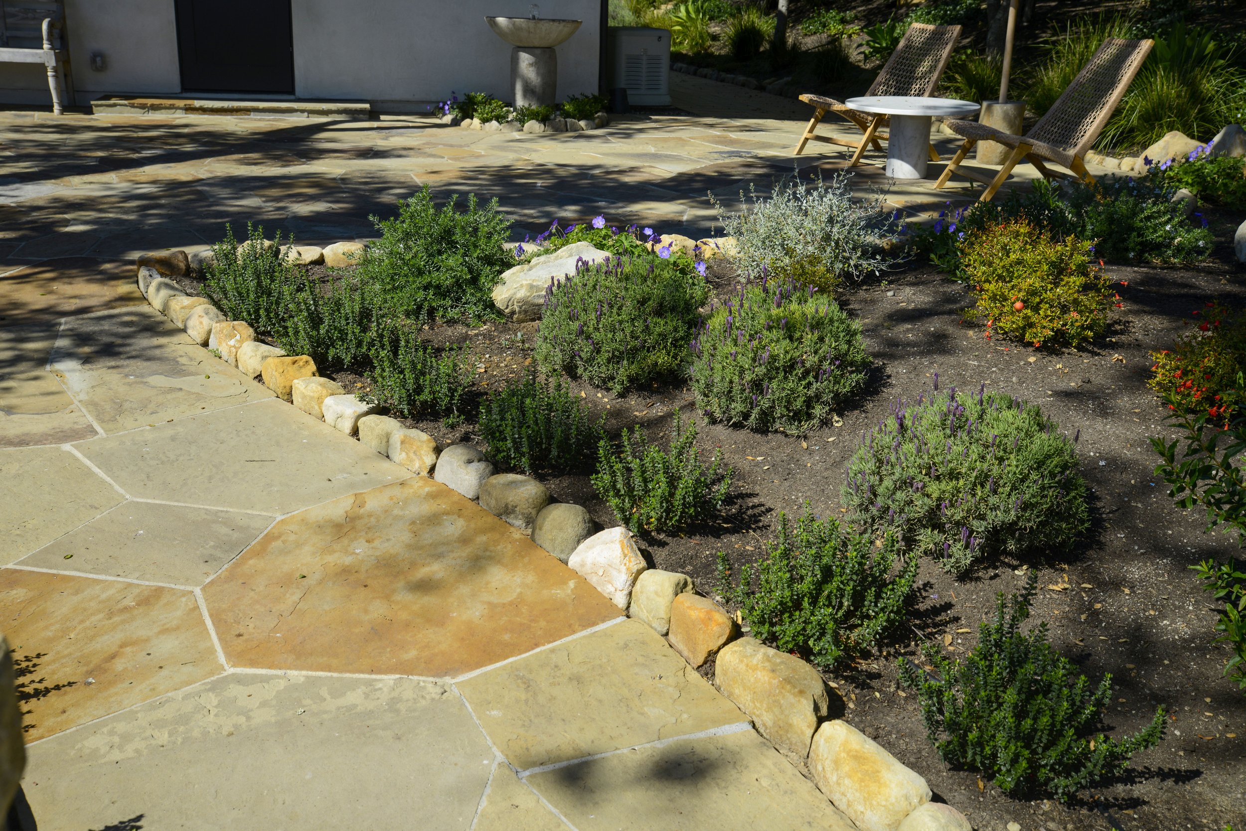 A landscaped backyard garden with a stone patio, a flower bed with various green plants and purple flowers, and two wooden chairs with a small round table in between, shaded by trees.