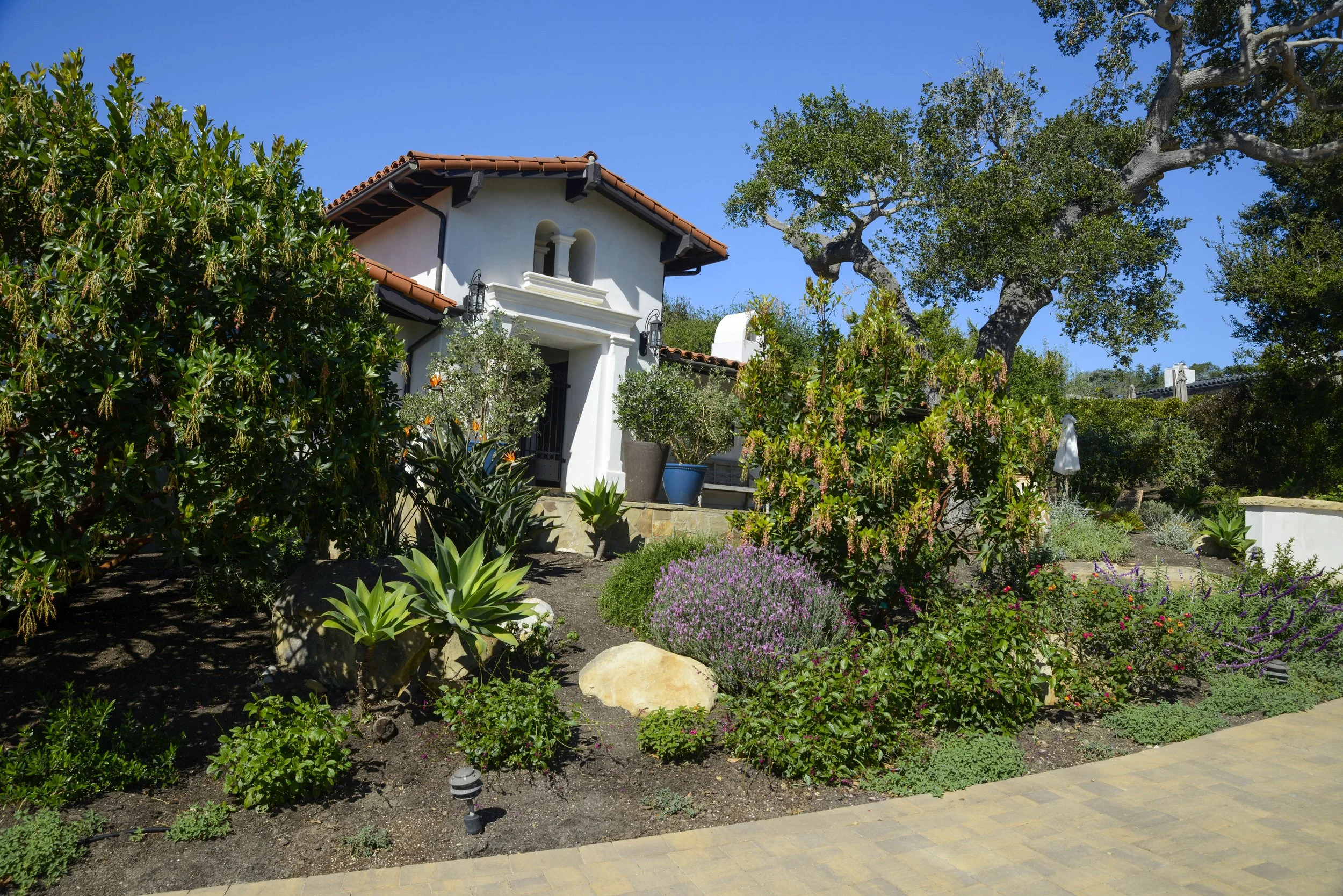 View of a house with a red tile roof surrounded by a lush, well-maintained garden with various plants and flowers, under a bright blue sky.