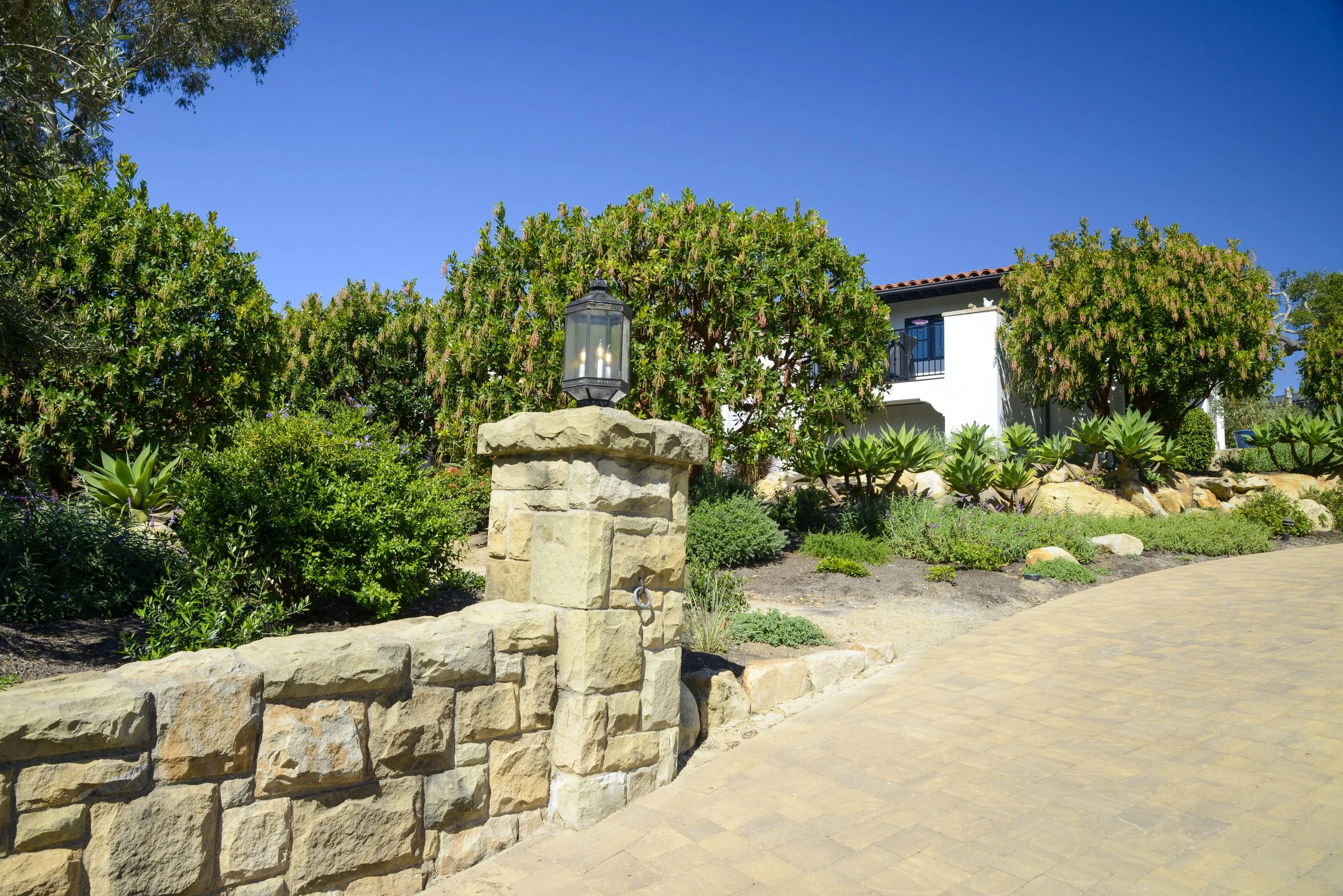 A stone wall with a lamp post on top, surrounded by lush green bushes and trees, with a white house and a paved walkway under a clear blue sky.
