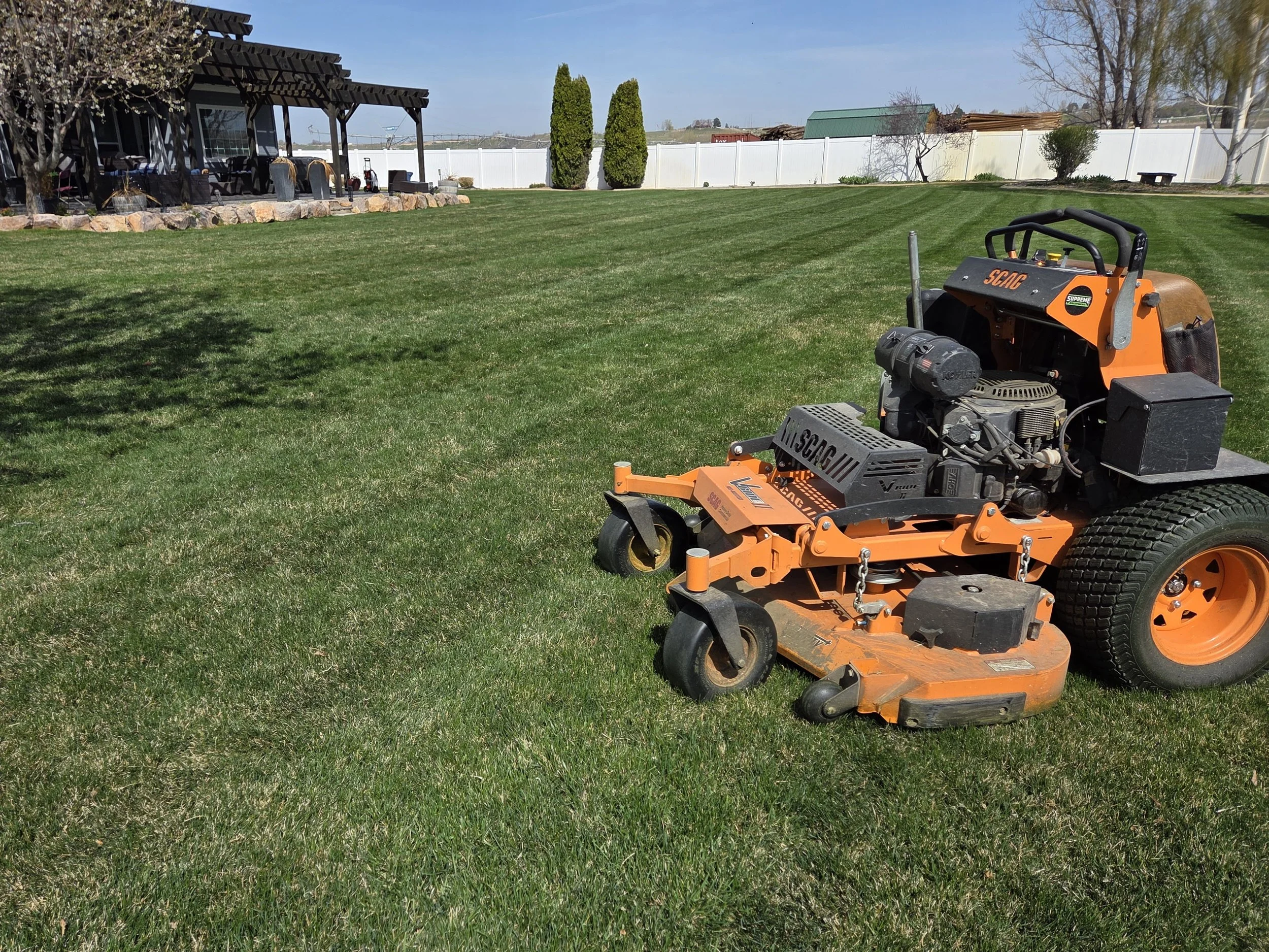 A lawn mower sitting on a well-maintained grassy backyard with a patio area, trees, and a white fence in the background.
