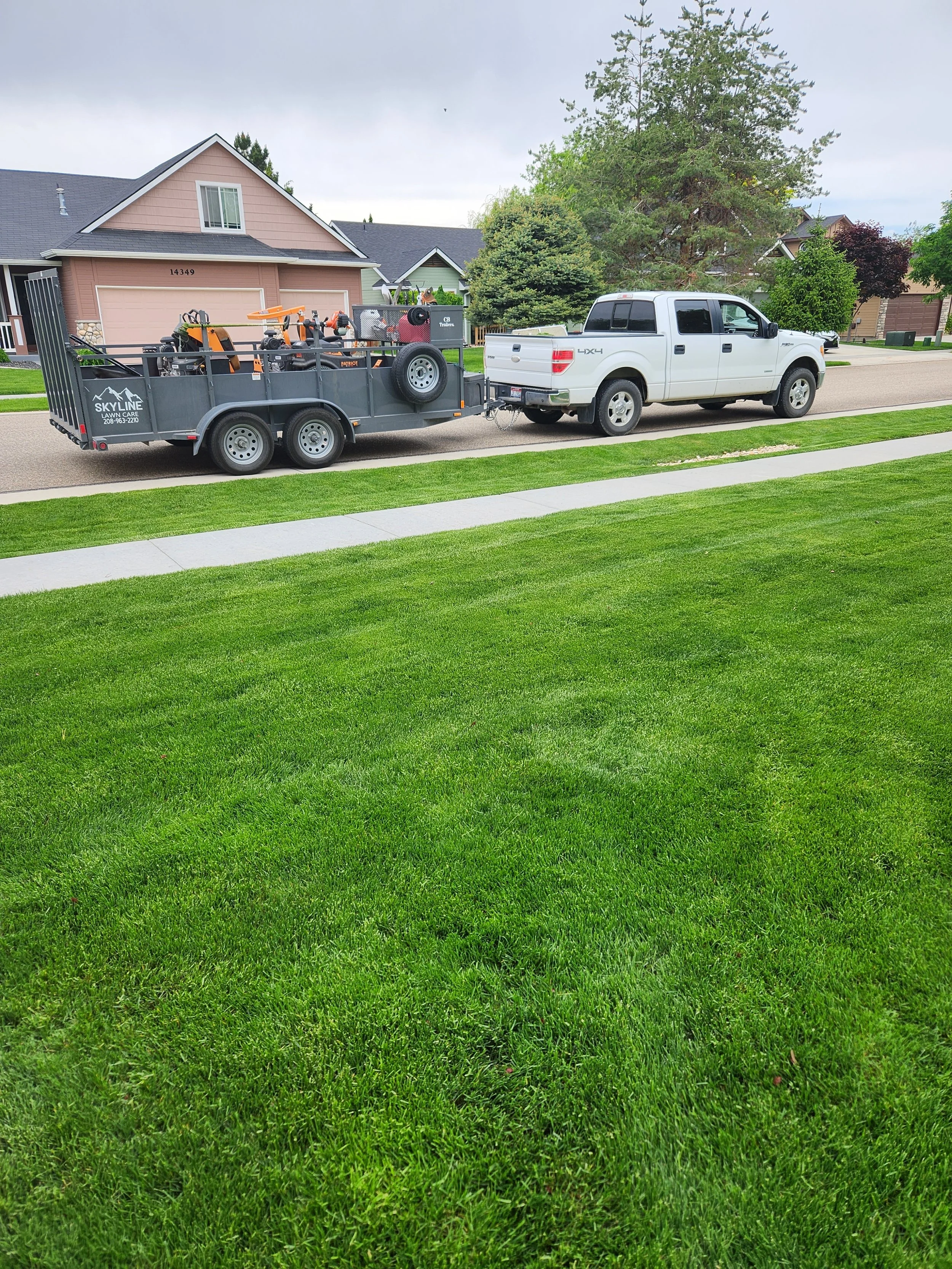 White pickup truck towing a trailer loaded with lawn care equipment on a suburban street.
