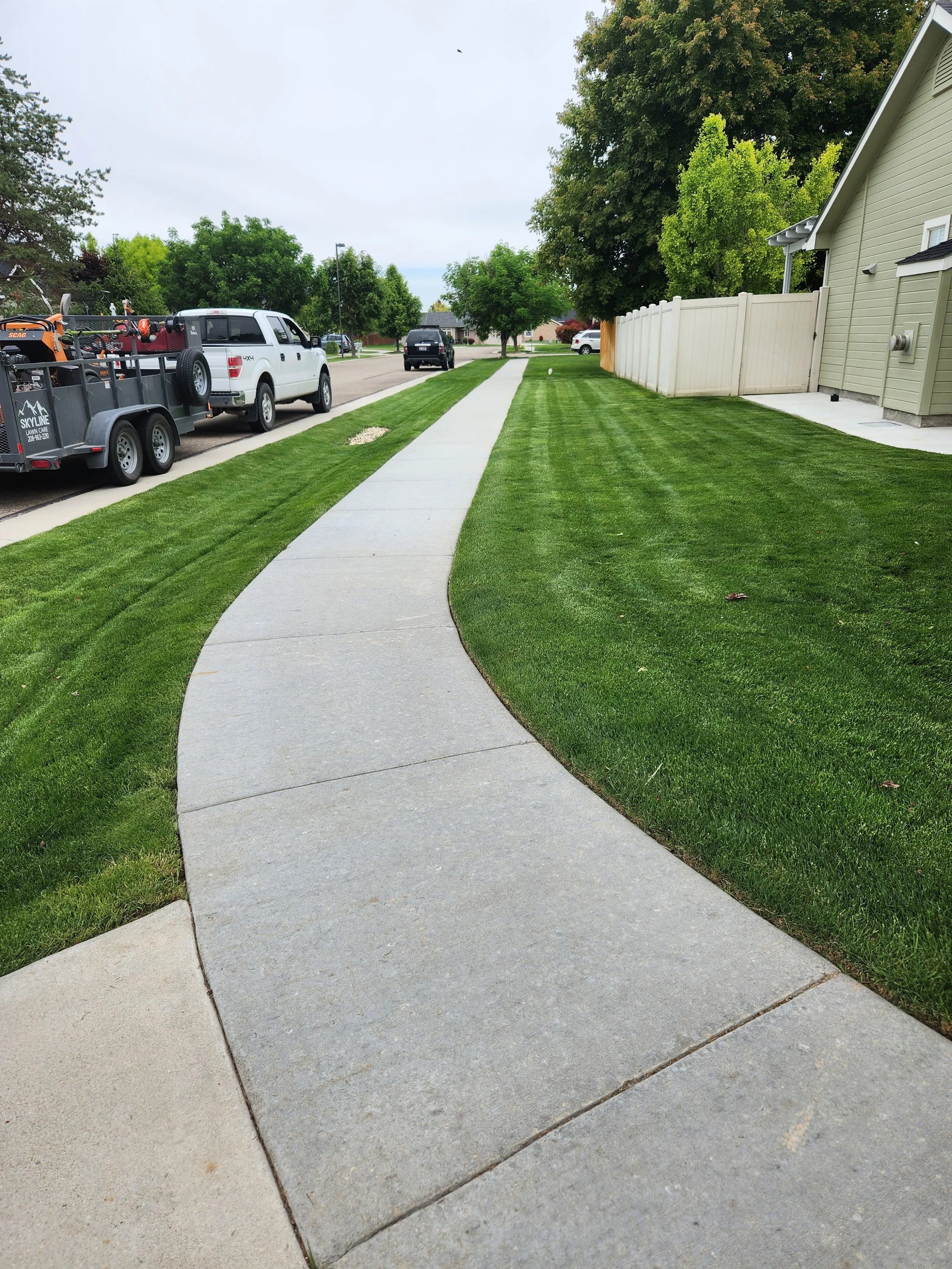 Curved concrete sidewalk running through well-maintained green lawn with trees and houses in the background.