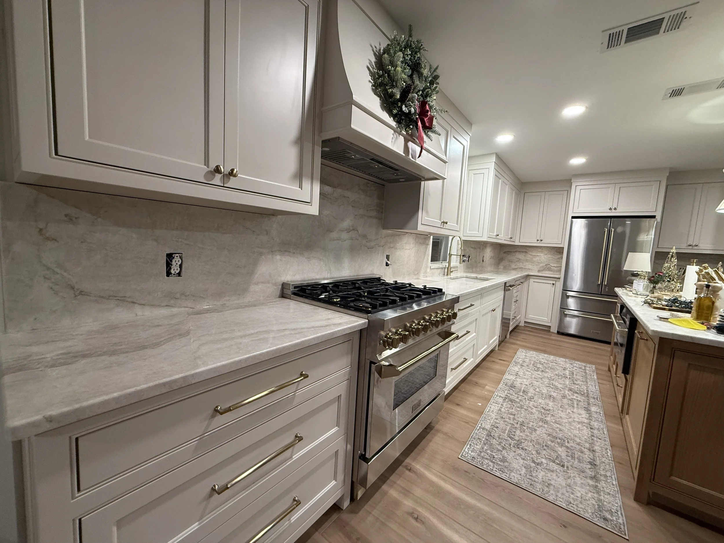 A modern kitchen with white cabinets, a marble countertop, stainless steel appliances including a refrigerator and oven, and a small decorated Christmas wreath hanging above the stove.