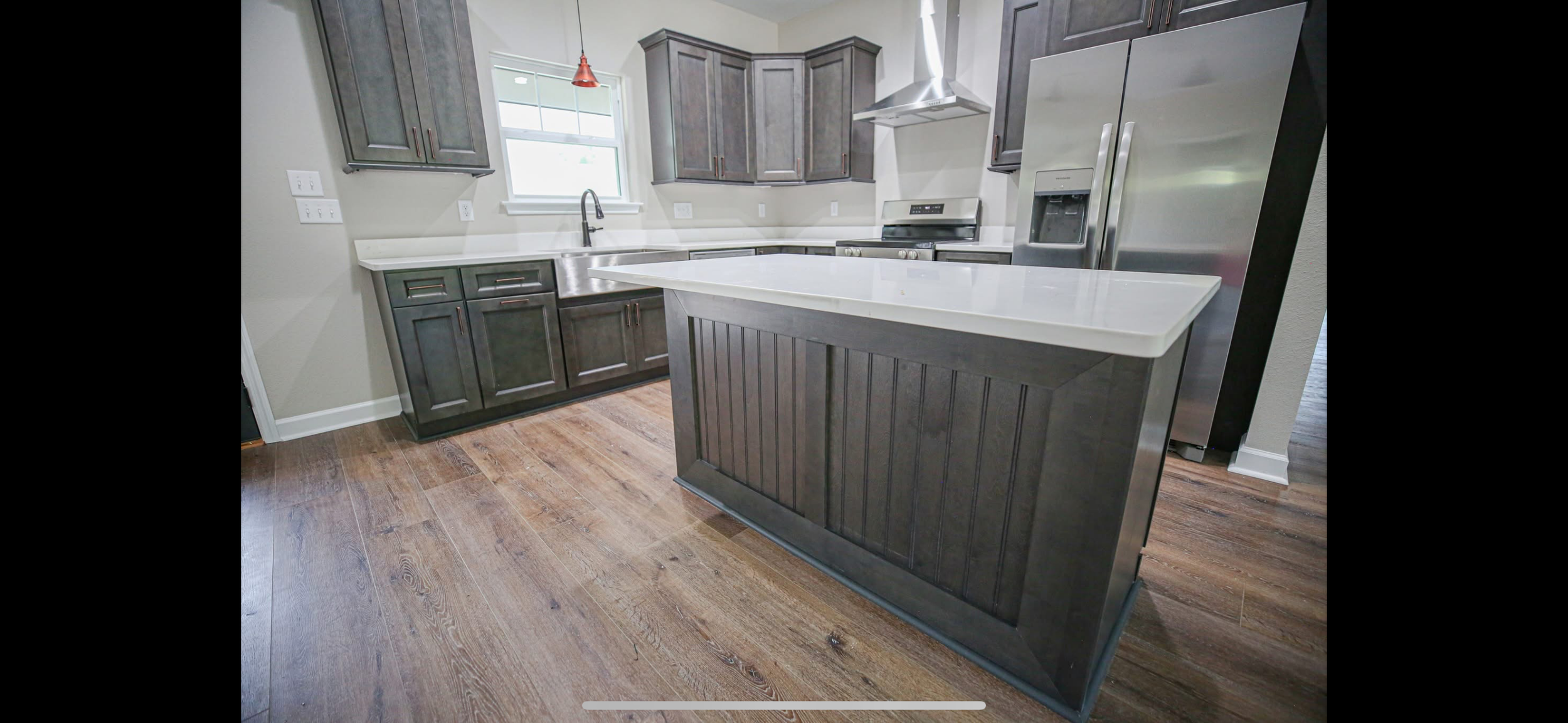Modern kitchen with dark gray cabinets, a stainless steel refrigerator, a white countertop island, and wood flooring.