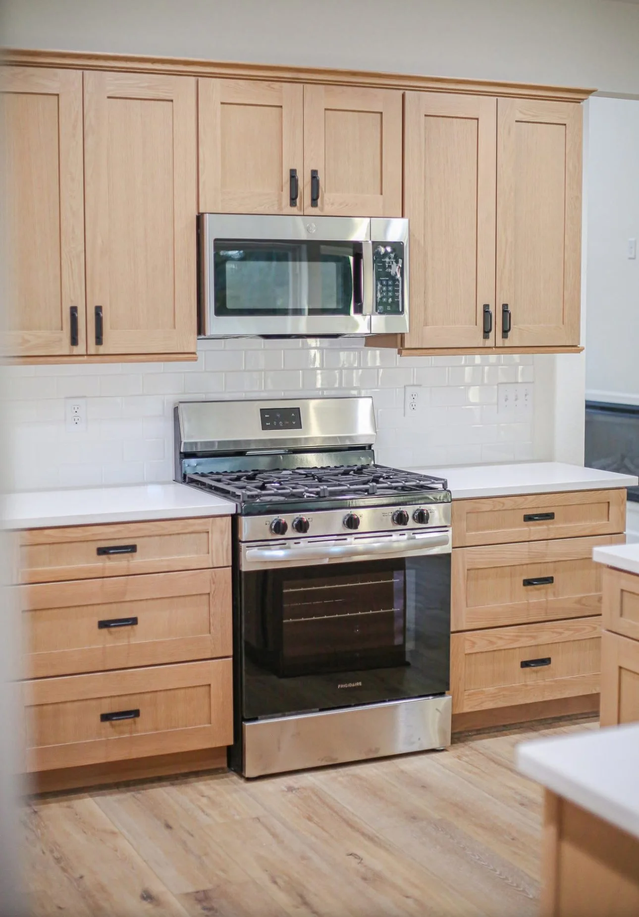 A modern kitchen with light wood cabinets, stainless steel appliances including a microwave above a gas stove, white countertops, and a light-colored wood floor.