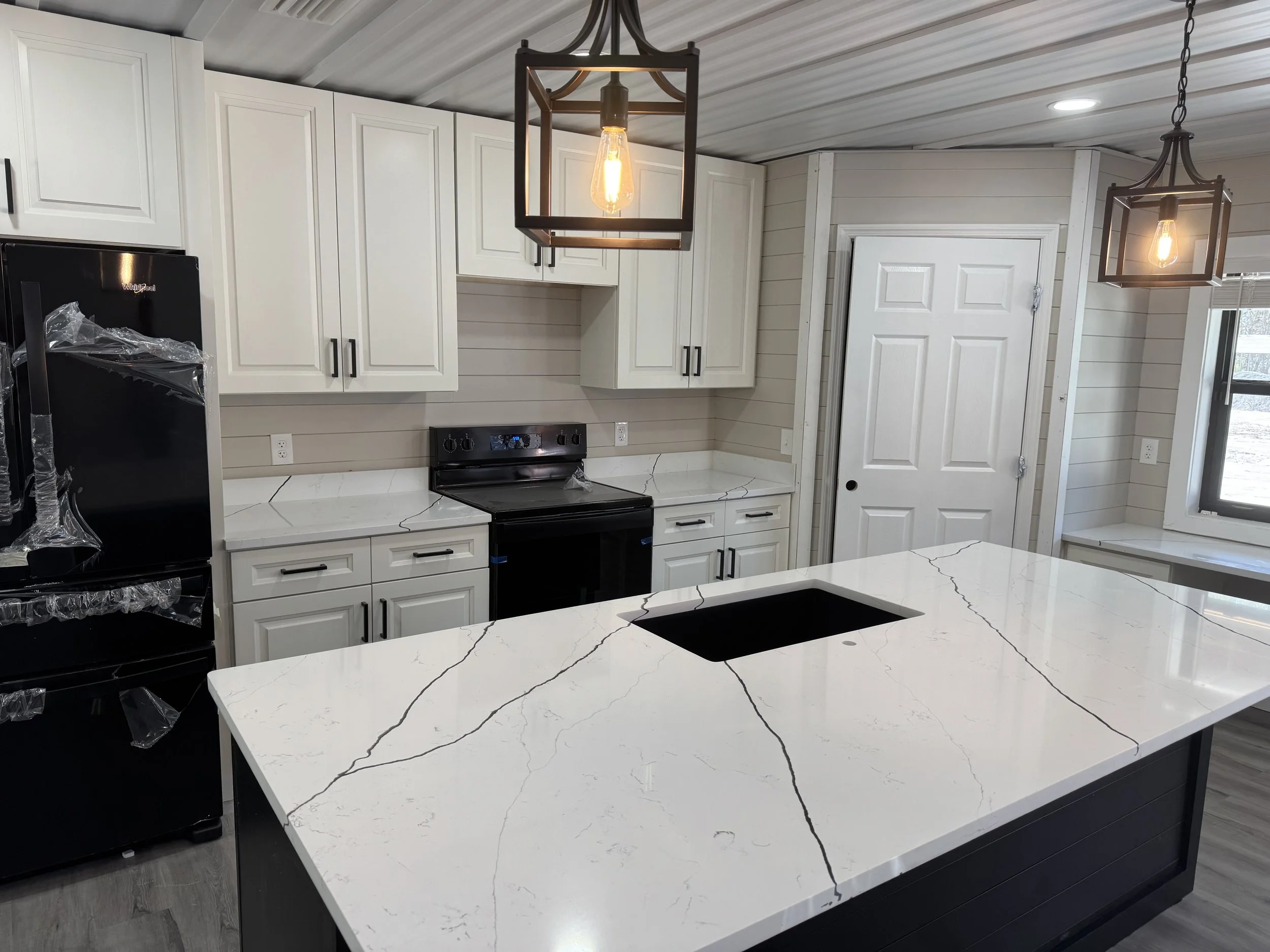 Modern kitchen with white cabinets, black appliances, marble island with black sink, pendant lights, window, and a door.