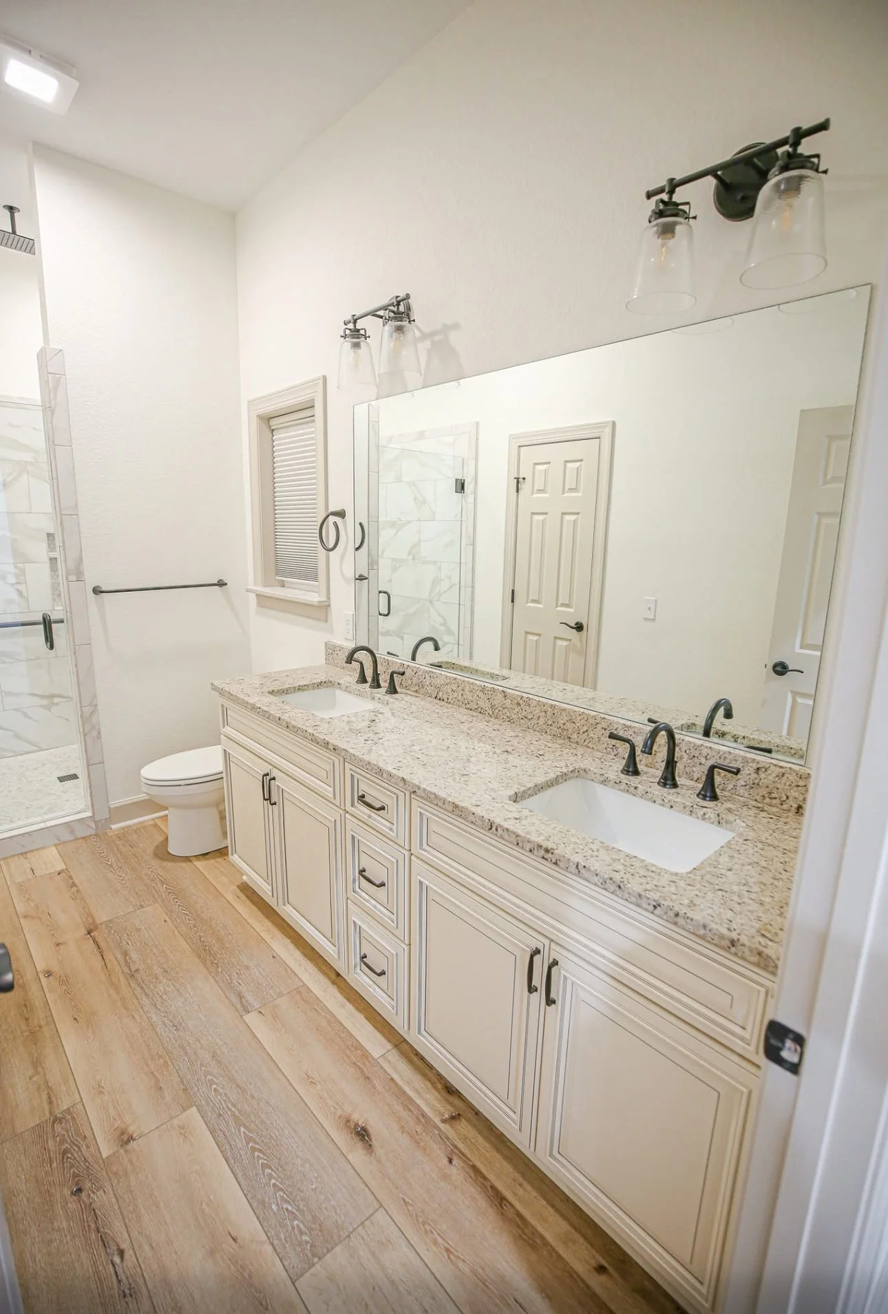 Bathroom with double vanity, granite countertop, oval sinks, black fixtures, large mirror, wood flooring, shower with glass door, and white walls.