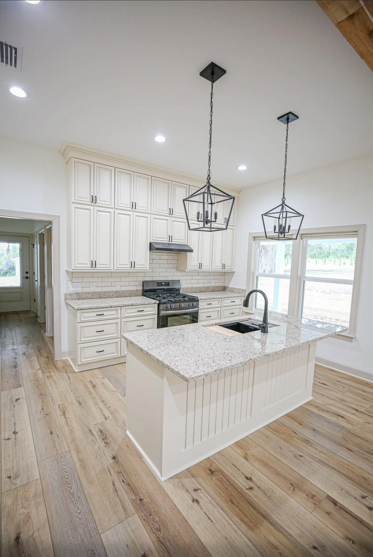 Bright kitchen with white cabinets, granite countertops, and a large island with a sink. Two black geometric pendant lights hang over the island. Light wood flooring and large windows are visible.