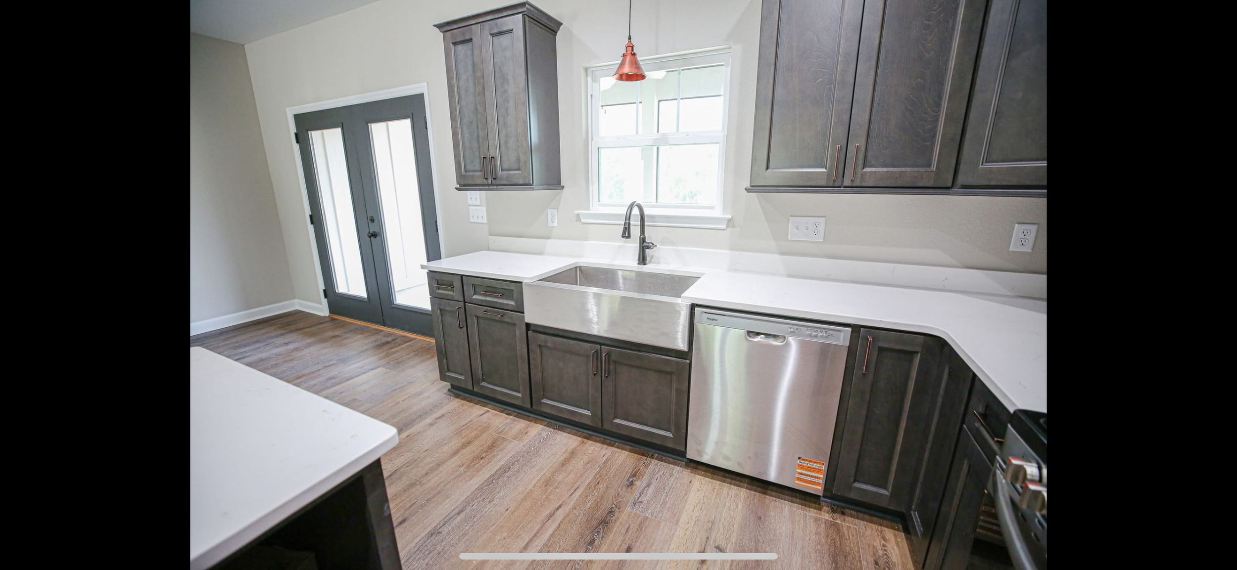 Modern kitchen with dark gray cabinets, white countertops, a farmhouse sink, stainless steel dishwasher, and hardwood floors. There is a window above the sink and a door with glass panels leading outside.