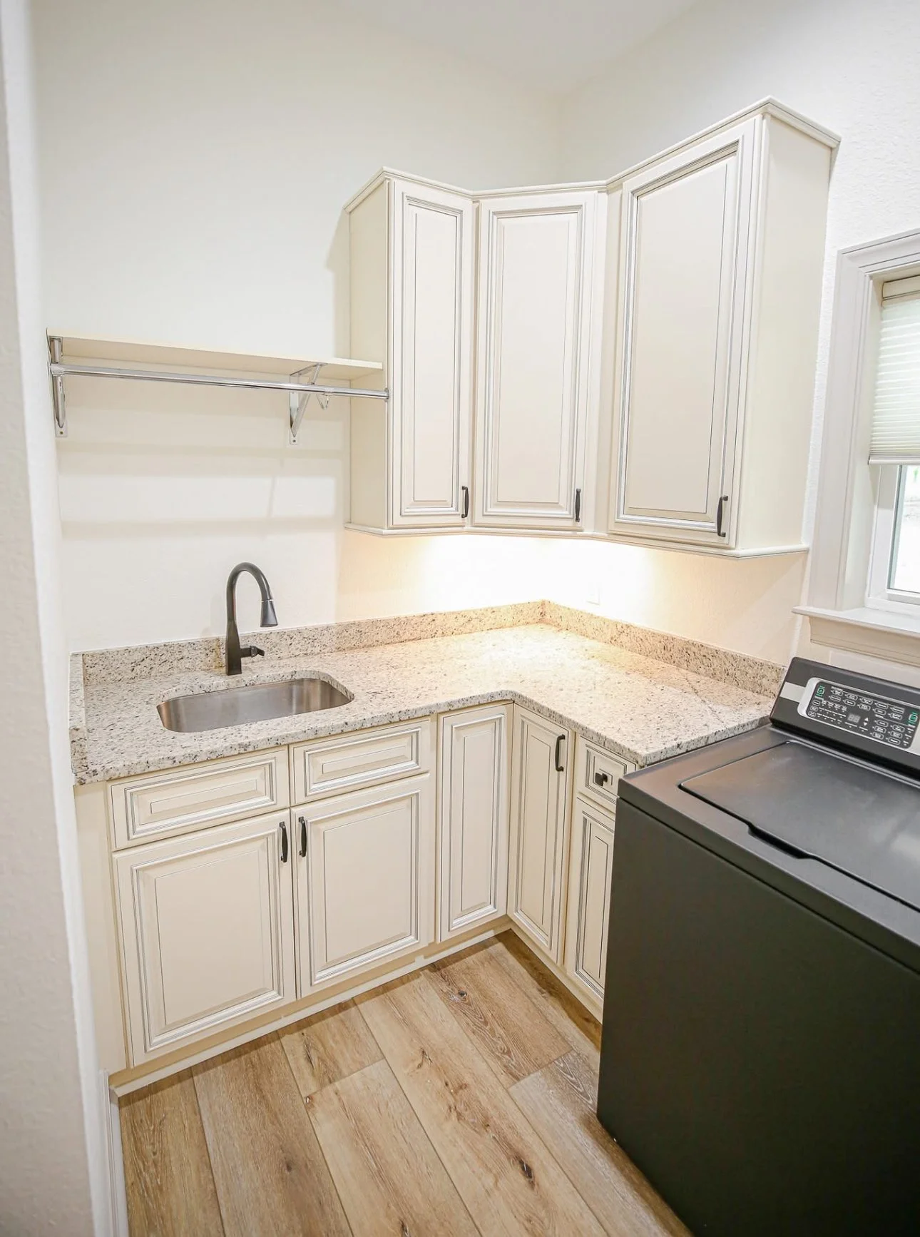 Kitchen with white cabinetry, granite countertops, a small sink, a black washing machine, and a window with blind, with light-colored wood flooring.