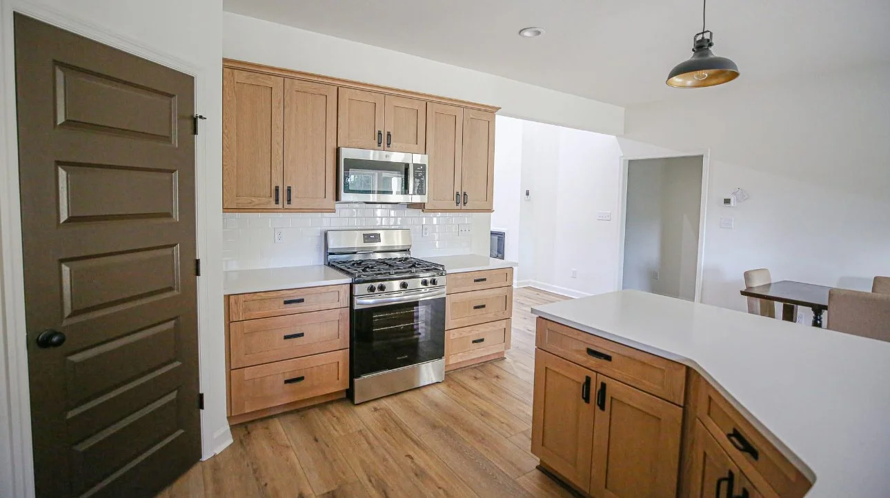 Kitchen with wooden cabinets, stainless steel stove and microwave, white quartz countertops, a dark brown door, hardwood flooring, and a dining area with a table and chairs.