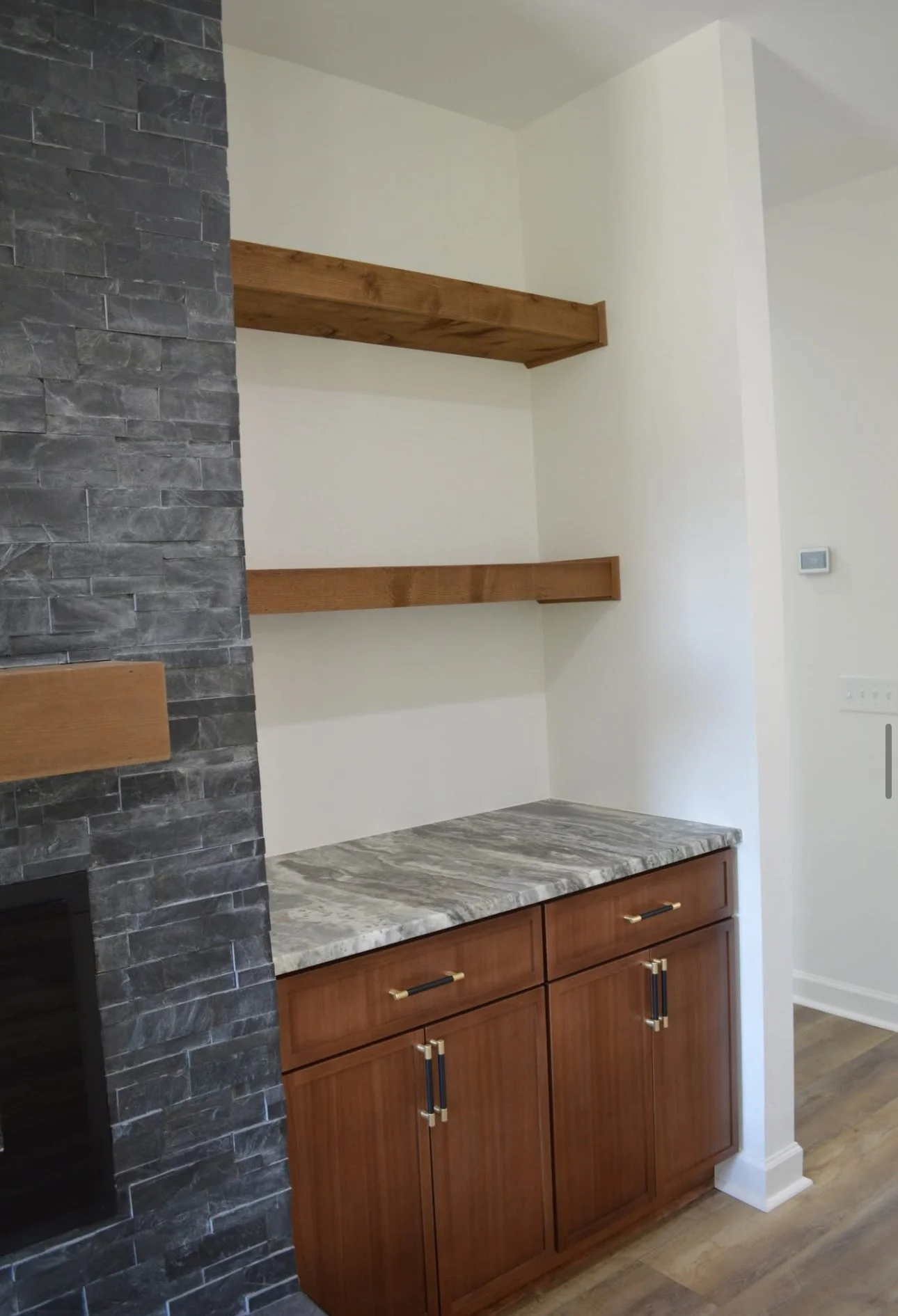 A section of a room with a countertop, wooden cabinets, and two wooden floating shelves on a white wall, next to a dark stone wall.