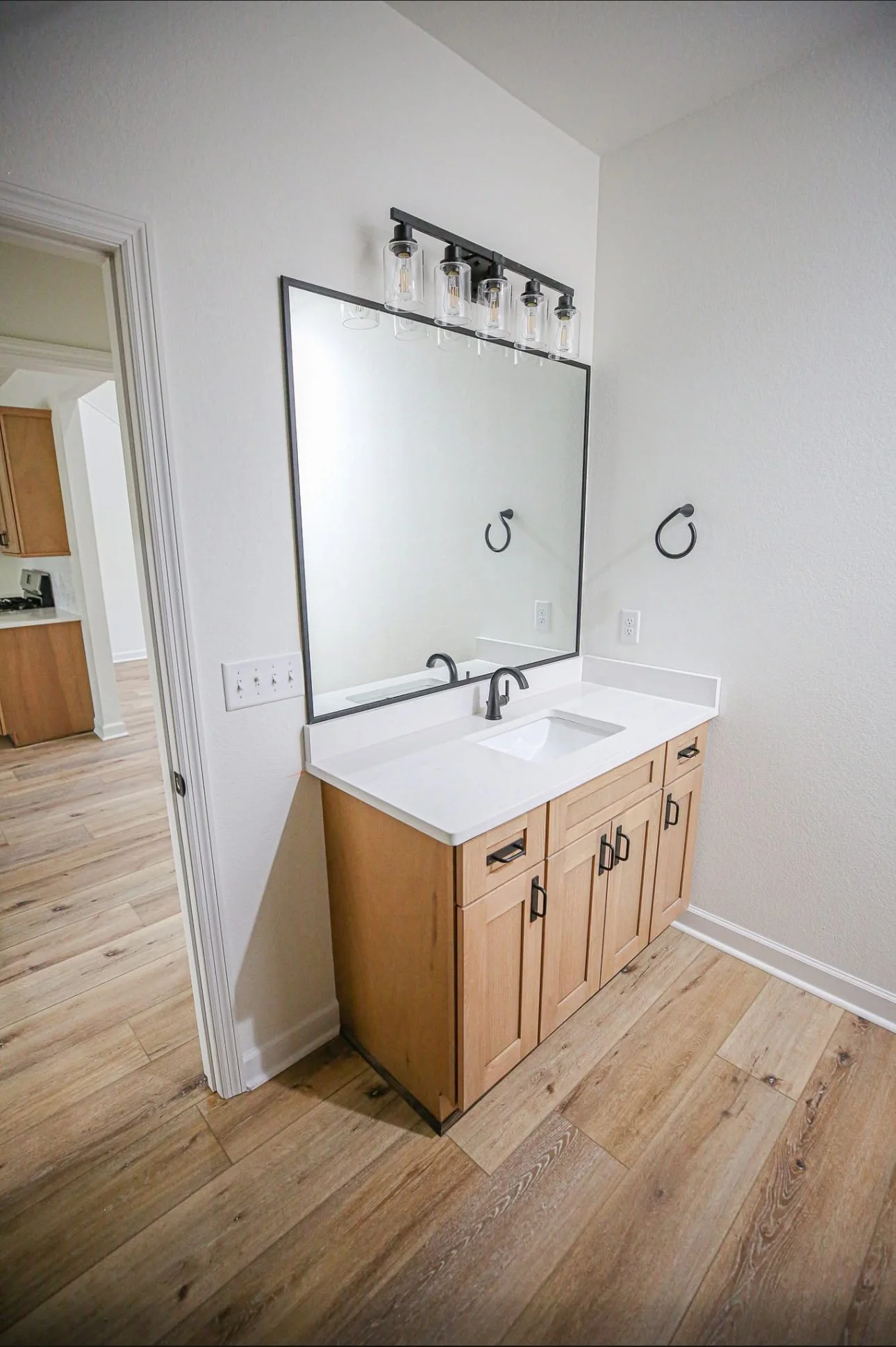 Bathroom vanity with a wooden cabinet, white countertop, rectangular undermount sink, large mirror, black wall-mounted tap, and black towel rings in a room with wooden flooring and white walls.
