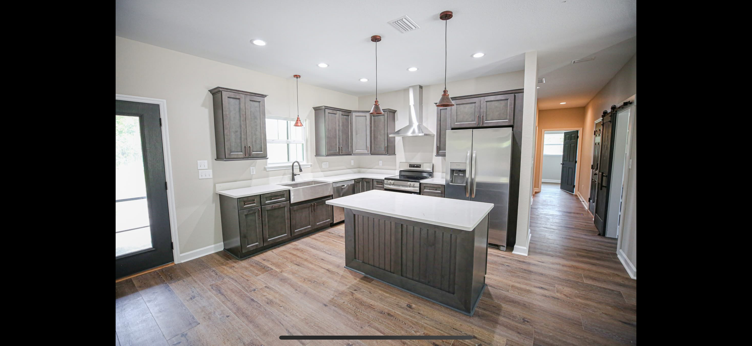 Modern kitchen with gray cabinets, white countertops, stainless steel refrigerator, stove, and vent hood, wood flooring, pendant lights, and a window above the sink.