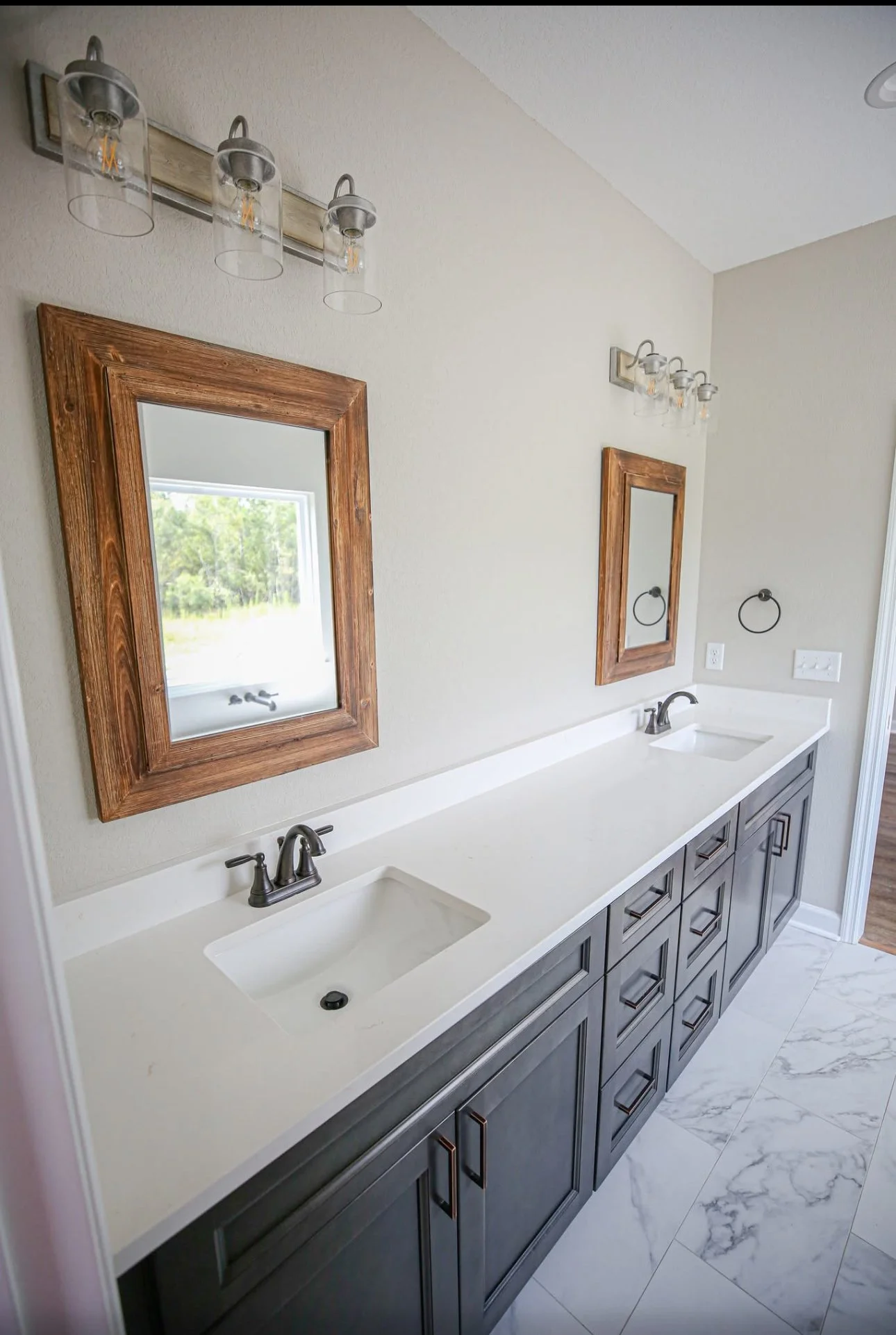 A modern bathroom with double vanity, two mirrors with wooden frames, three-lamp light fixtures, and marble floor tiles.