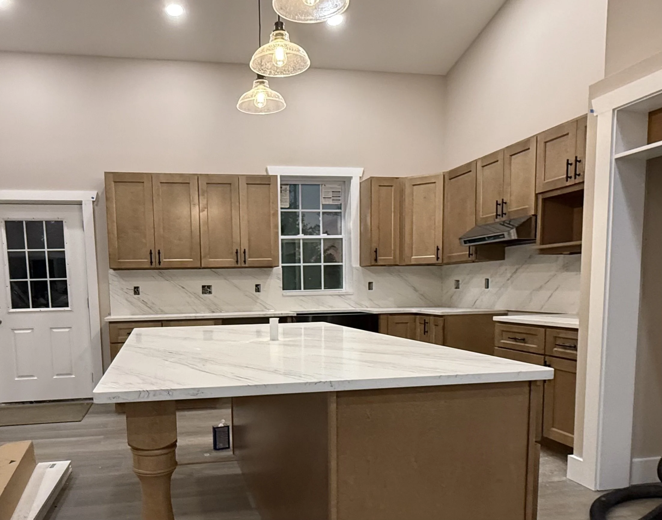 A modern kitchen with wooden cabinets, white marble countertops, a central island, and a window above the sink. There are three hanging light fixtures and a door on the left.