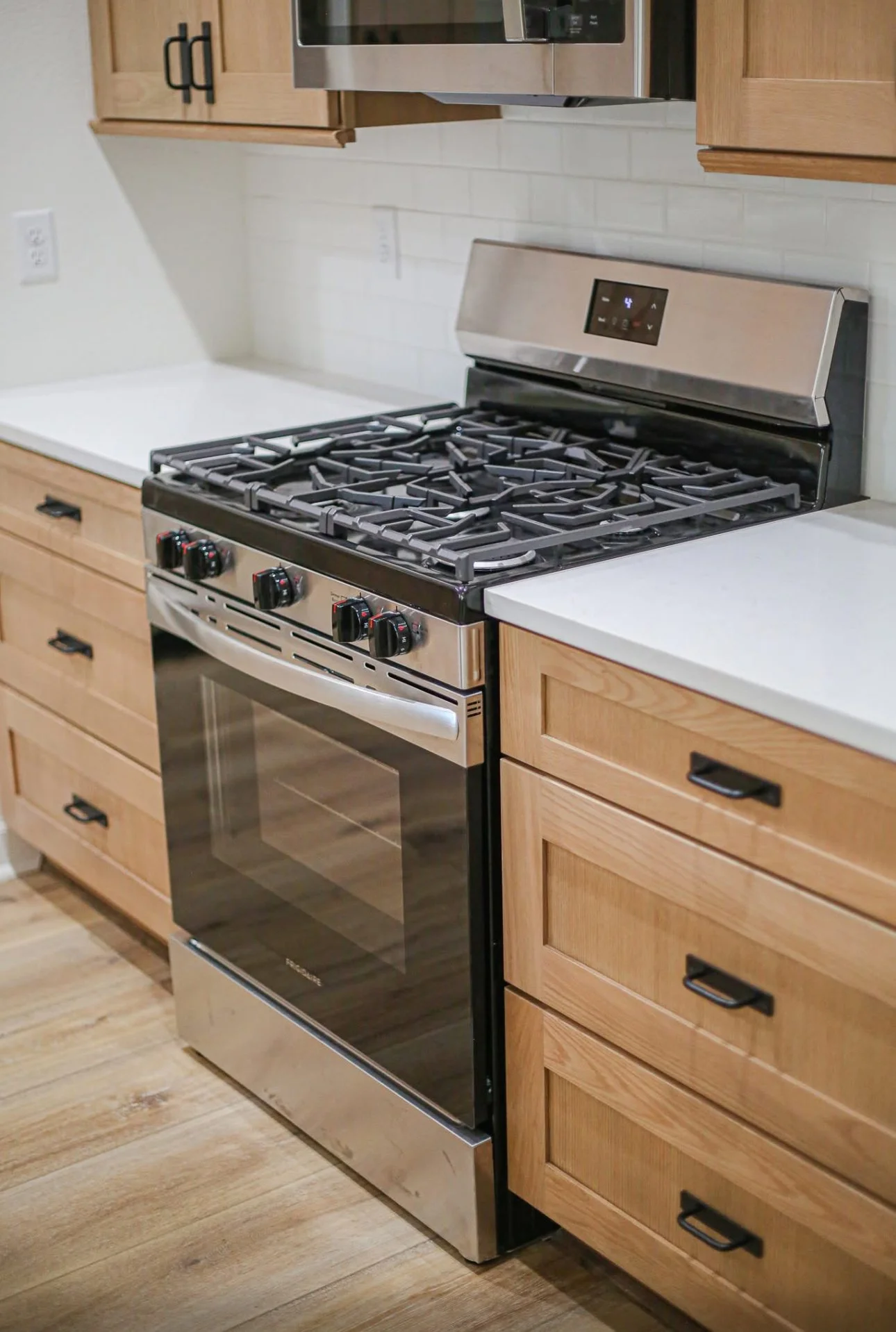 A stainless steel gas stove with four burners, black control knobs, and an oven, surrounded by wooden kitchen cabinets with black handles, white countertops, and a microwave above.