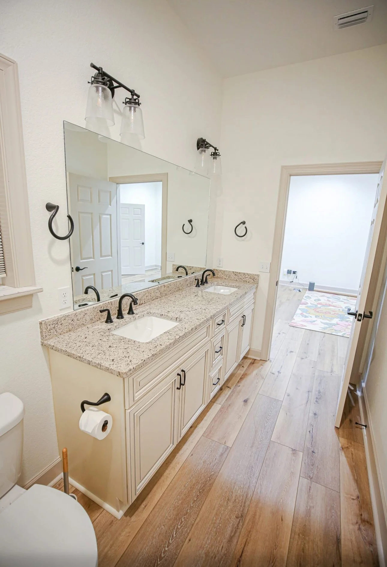 Bathroom with a dual sink vanity with granite countertops, large mirror, black fixtures, light wood cabinets, and wall-mounted towel rings. A toilet is visible on the left, and decor in the adjacent room is partially visible through the open door.