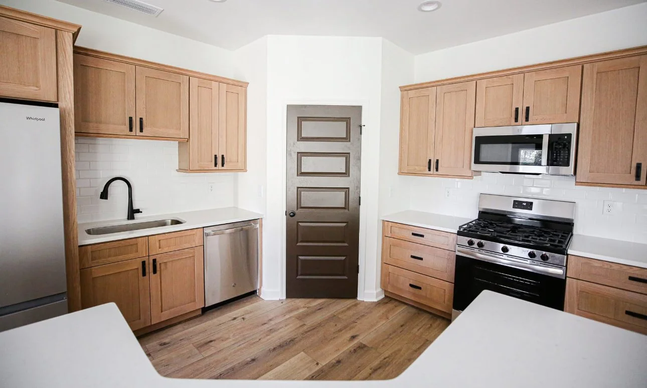 Kitchen with wooden cabinets, white countertops, stainless steel appliances including a refrigerator, dishwasher, oven, and microwave, and a wooden floor. A dark door is centered on the back wall.