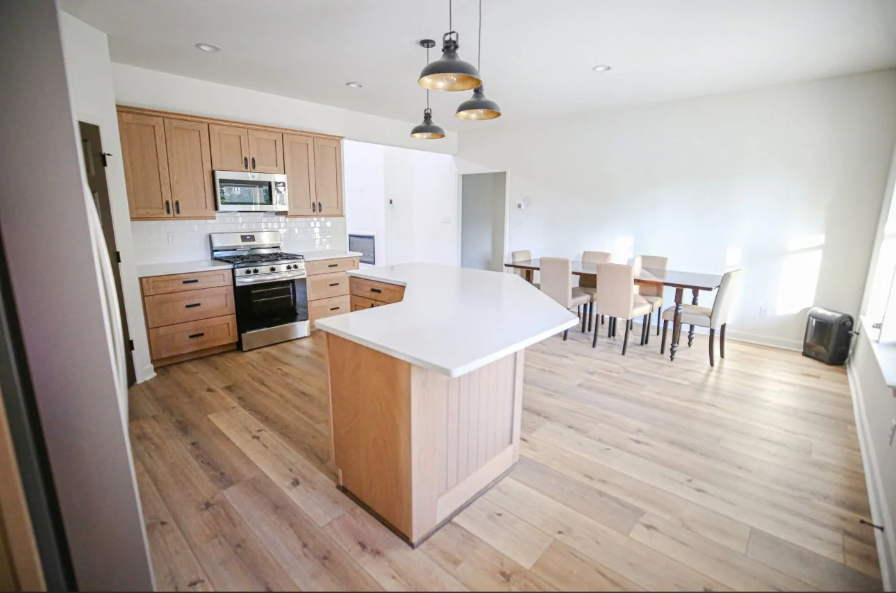 Photo of a bright, modern kitchen and dining area with light wood cabinets, a kitchen island with a white countertop, a stainless steel stove and microwave, a dining table with eight beige chairs, and hardwood flooring.