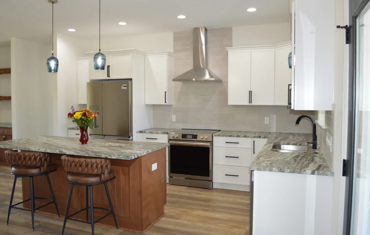Modern kitchen with white cabinets, stainless steel appliances, granite countertops, a kitchen island with a wood finish, and two bar stools. There are pendant lights hanging over the island, a vase with flowers, and a sliding glass door on the right