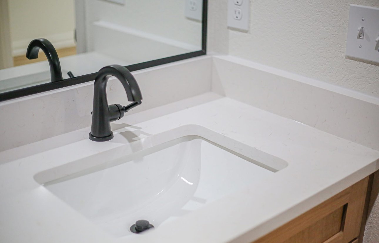 A bathroom sink with a black faucet, a mirror, two electrical outlets, and a light switch on the wall.