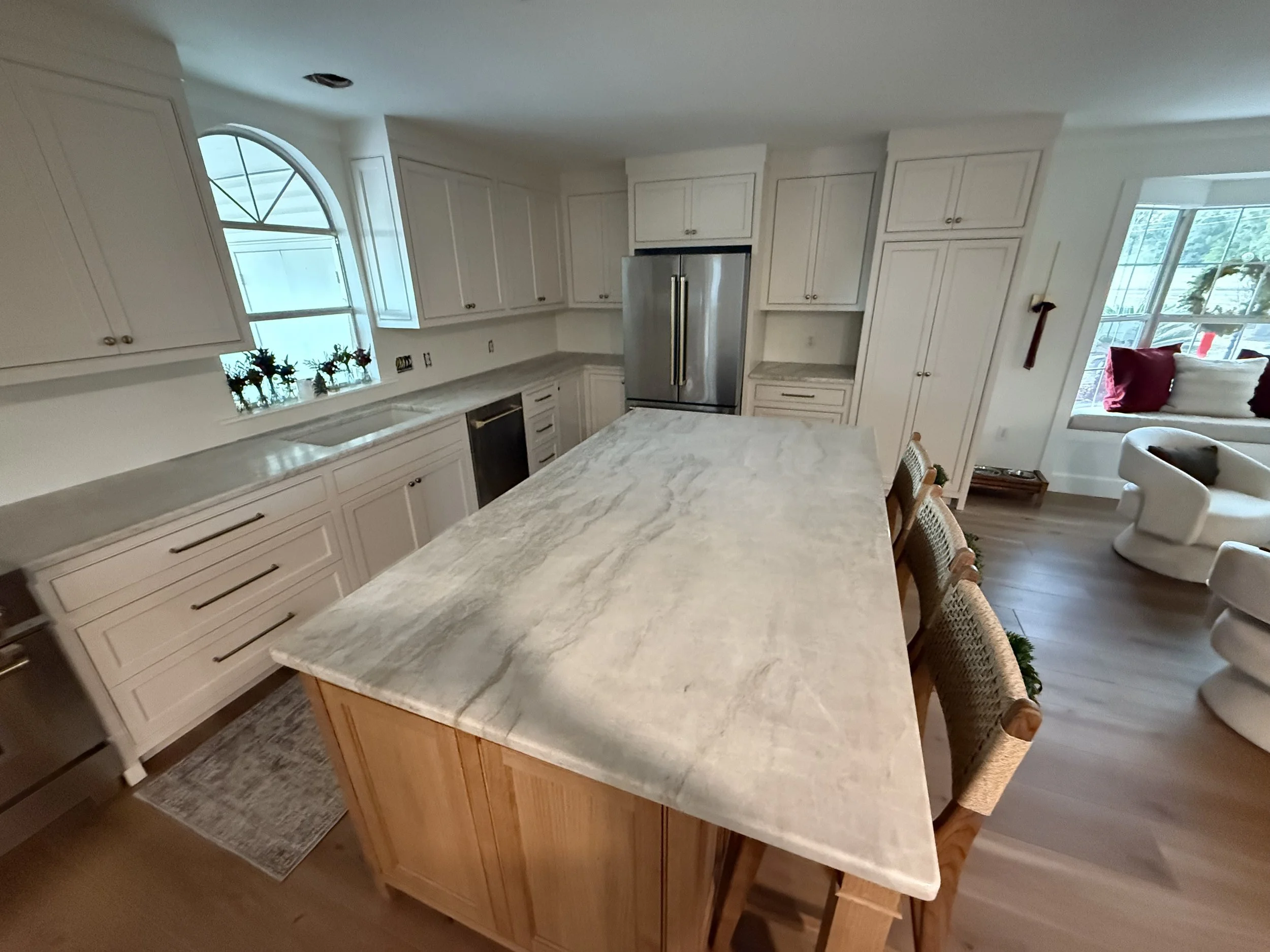 A spacious kitchen with white cabinets, a large marble island, stainless steel refrigerator, a window with flower pots, and a doorway leading to a sitting area with white furniture and red pillows.
