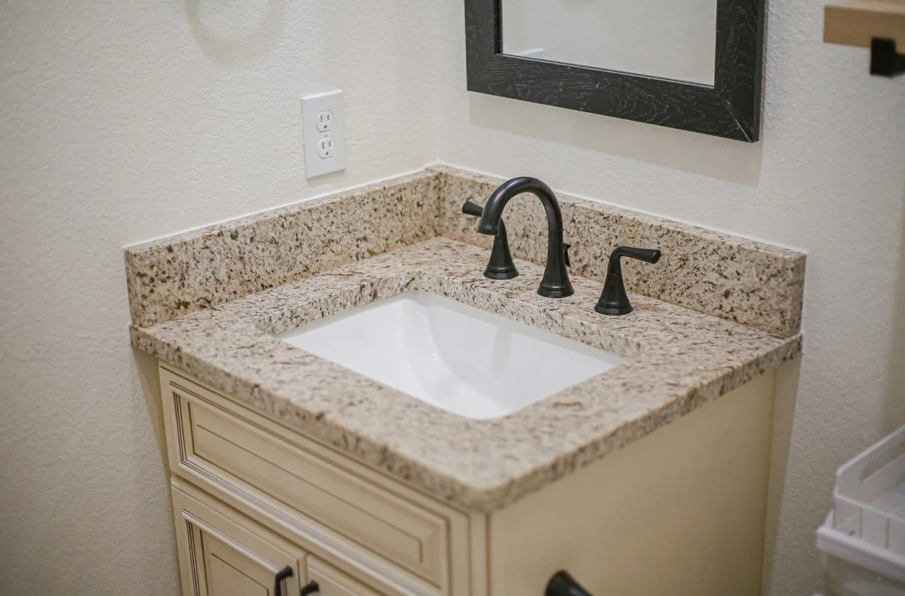 Bathroom sink with granite countertop, black faucet and handles, mirror, electrical outlet on beige wall, part of white cabinet below, and a plastic storage container to the right.