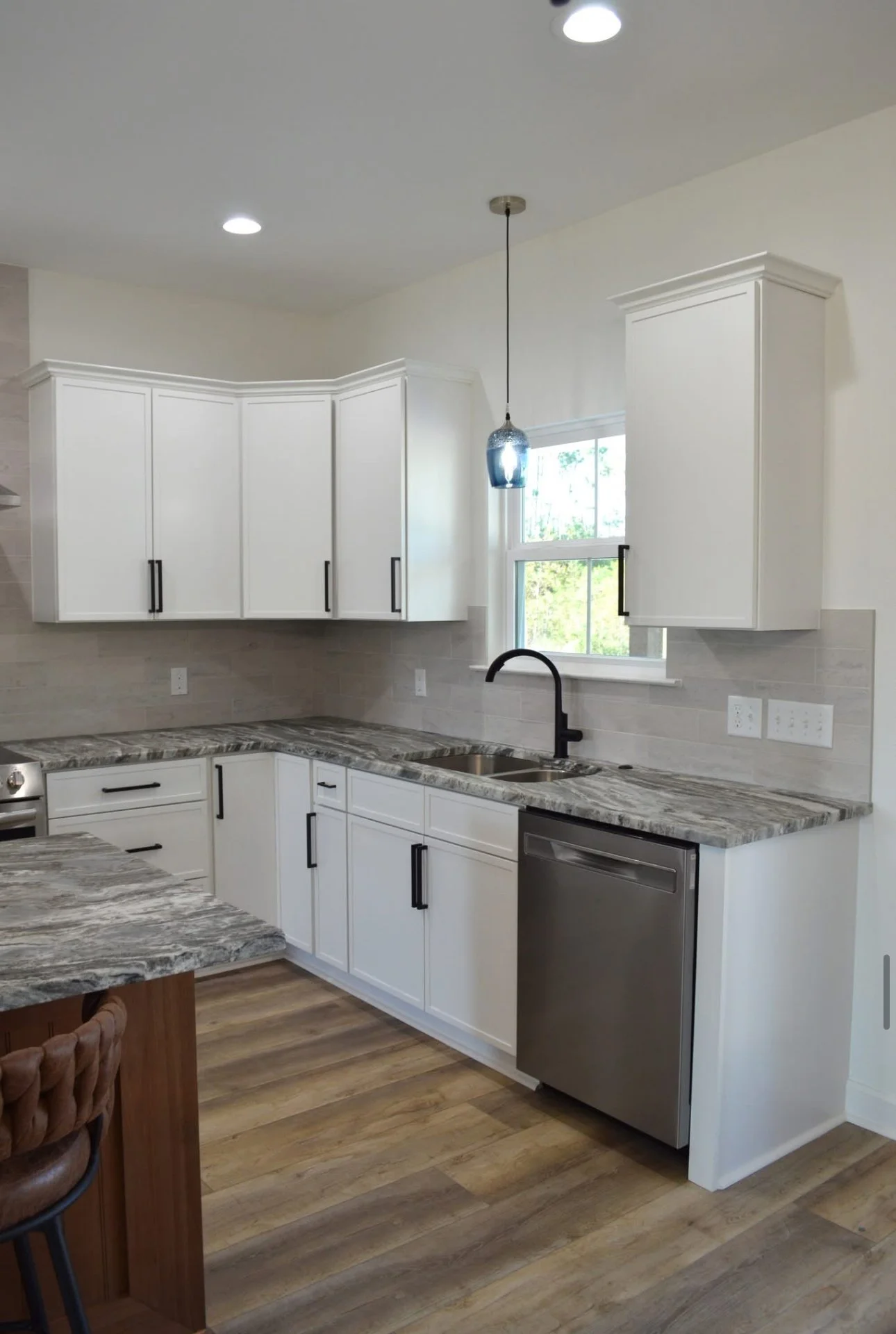 Modern kitchen with white cabinets, black handles, marble countertops, stainless steel dishwasher, and a black faucet, with a window above the sink and a pendant light.