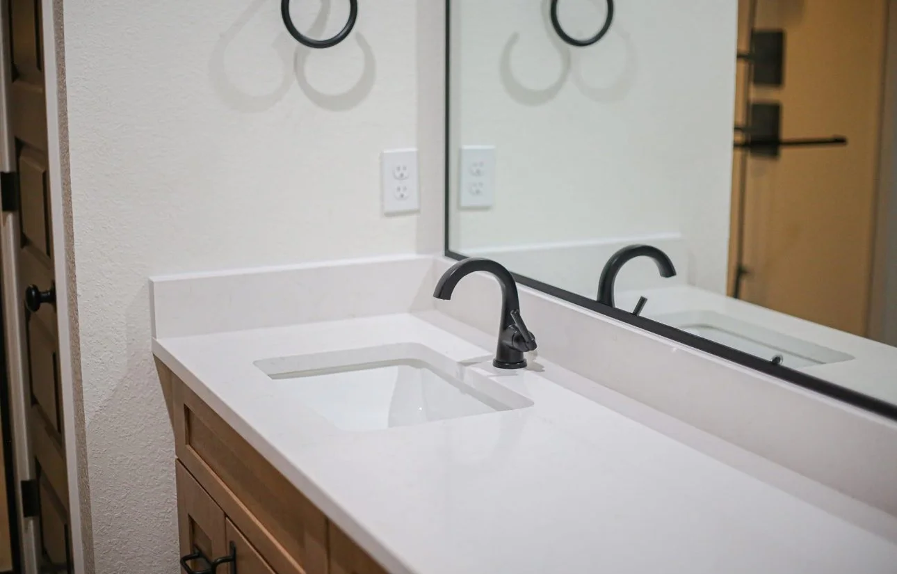 Bathroom vanity with a white countertop, black faucet, and a large mirror reflecting part of the scene and a door.