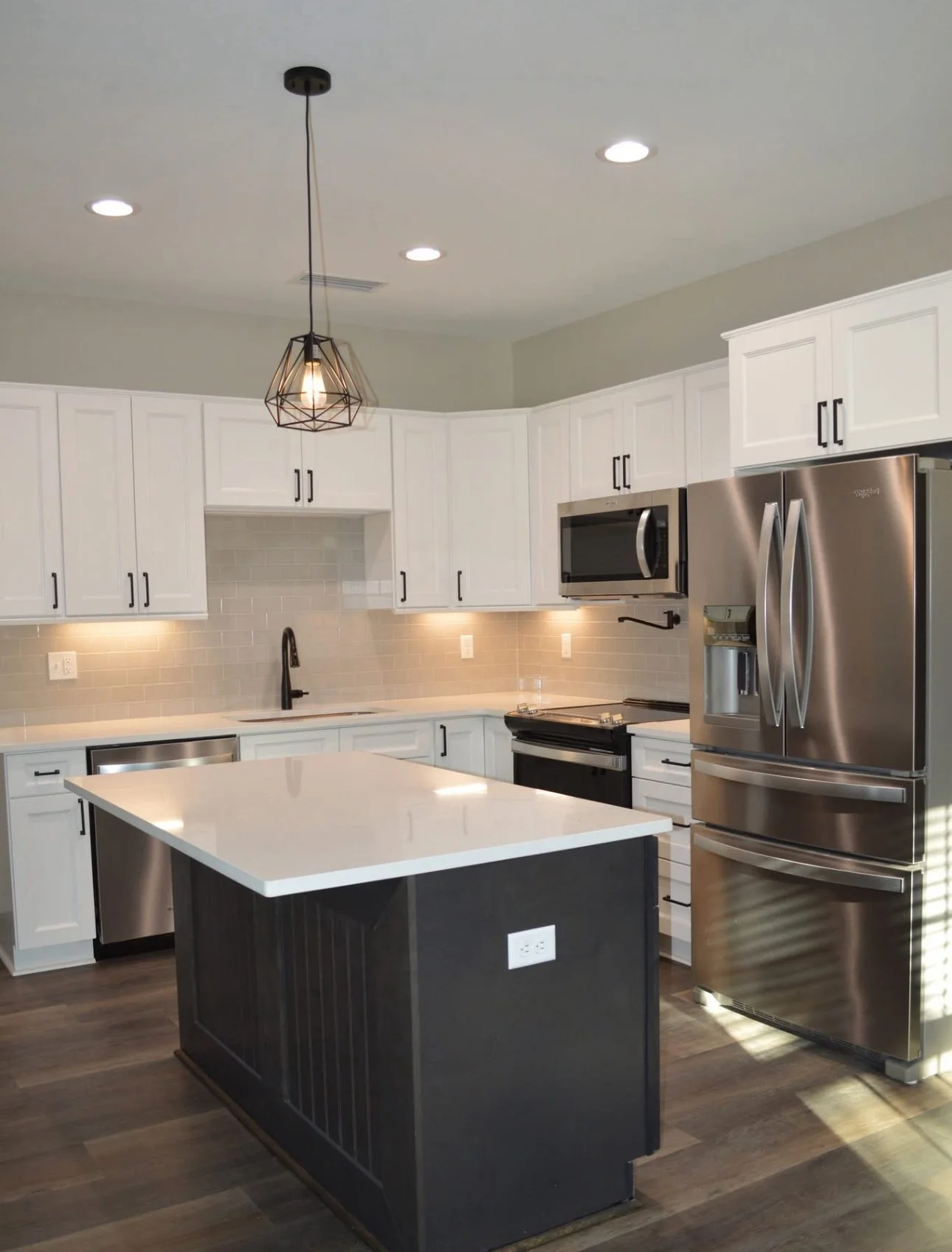 Modern kitchen with white cabinets, stainless steel appliances, dark island with electrical outlet, pendant light, and hardwood flooring.