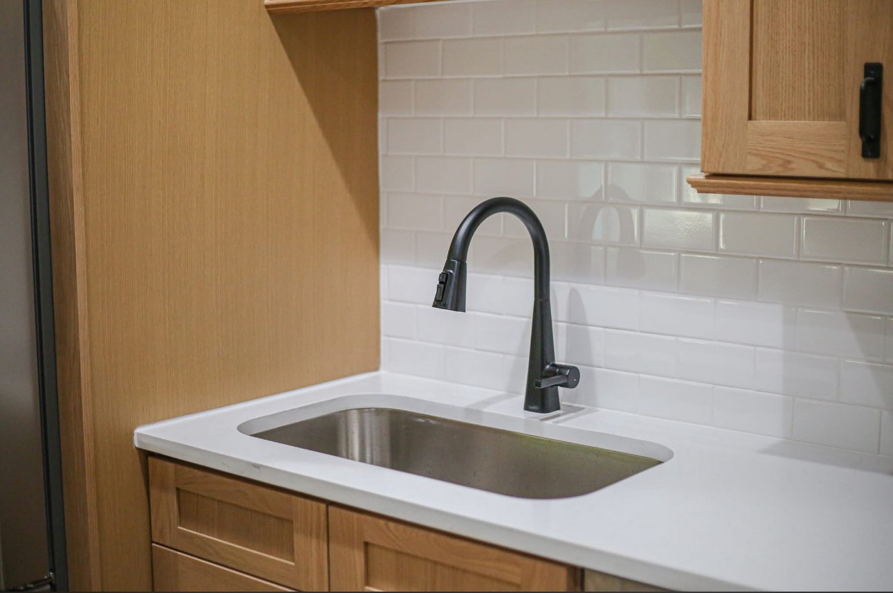 Kitchen sink with a black faucet, white countertop, wooden cabinets, and a white tiled backsplash.