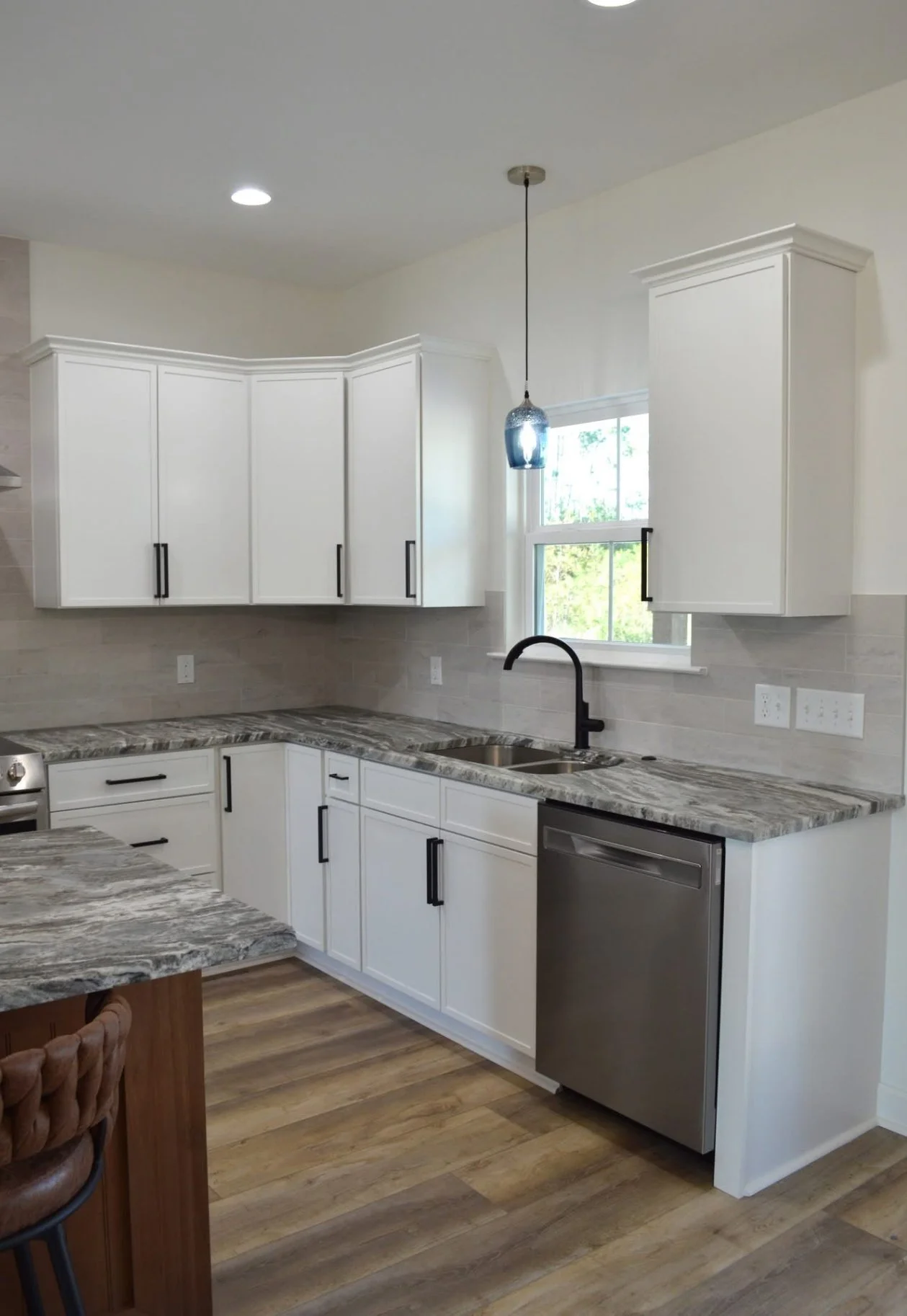 Modern kitchen with white cabinets, granite countertops, stainless steel dishwasher, black faucet, hanging pendant light, and wood flooring.