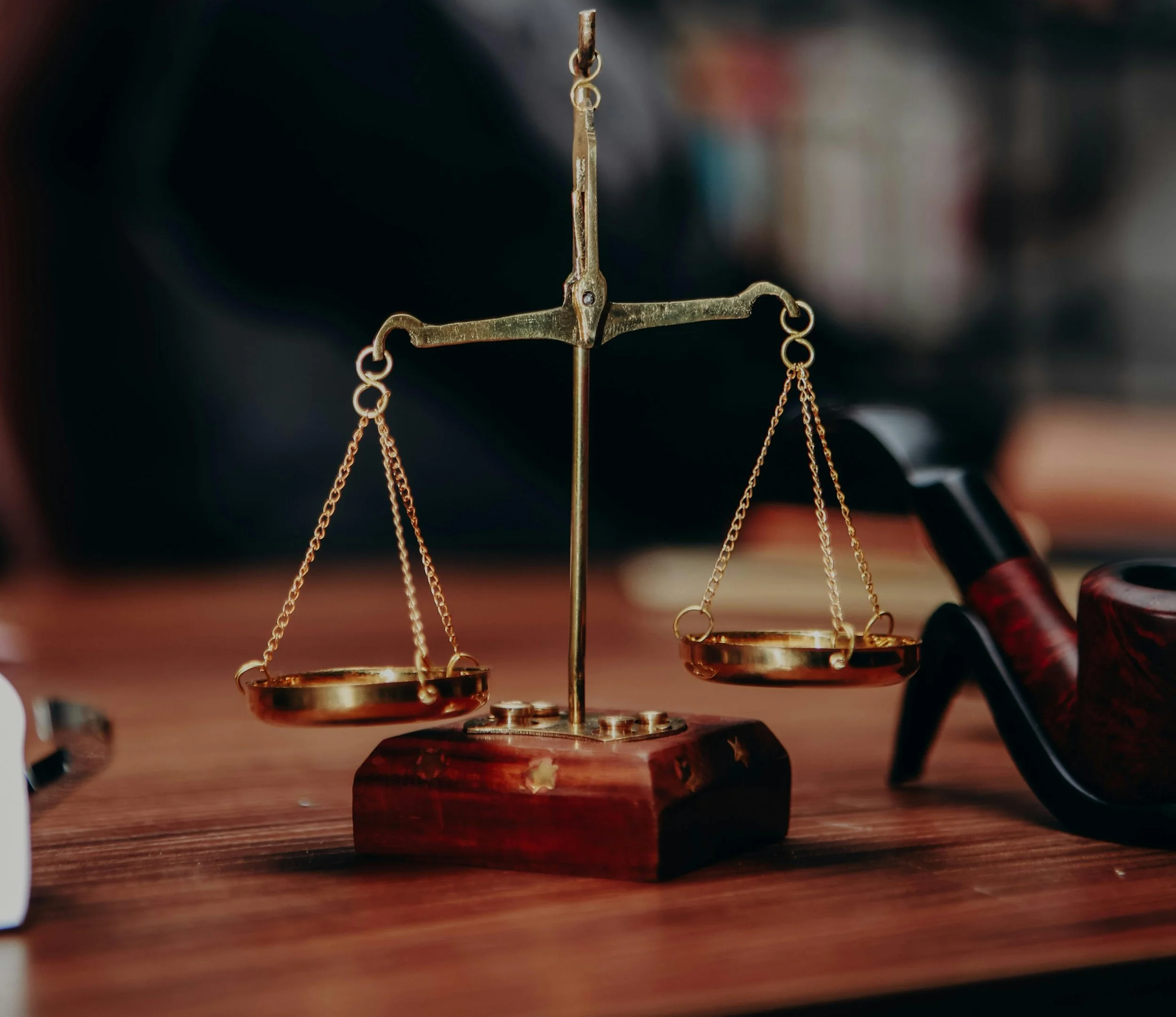 A set of brass balance scales placed on a wooden surface with scattered coins, and a dark wooden pipe is visible on the right.