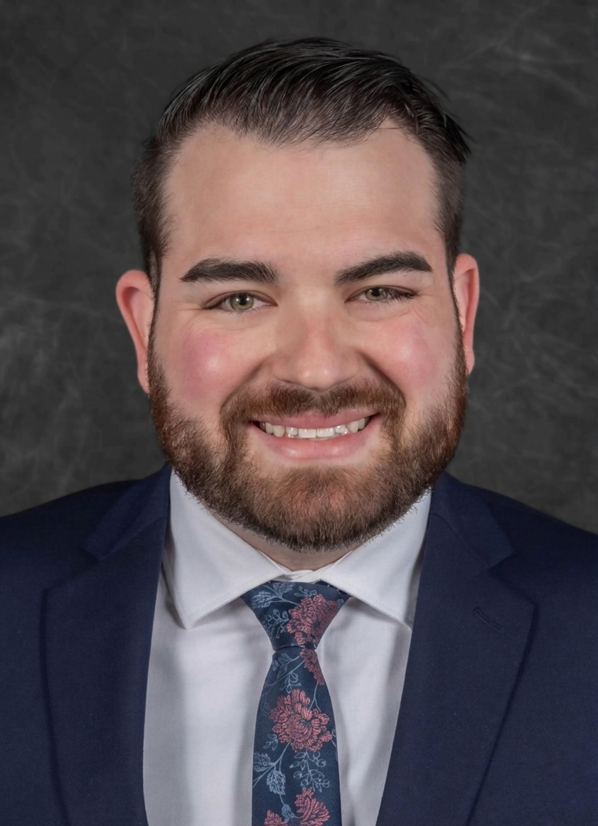 A smiling man with short dark hair, a beard, wearing a navy blue suit, white dress shirt, and a floral tie, against a dark textured background.