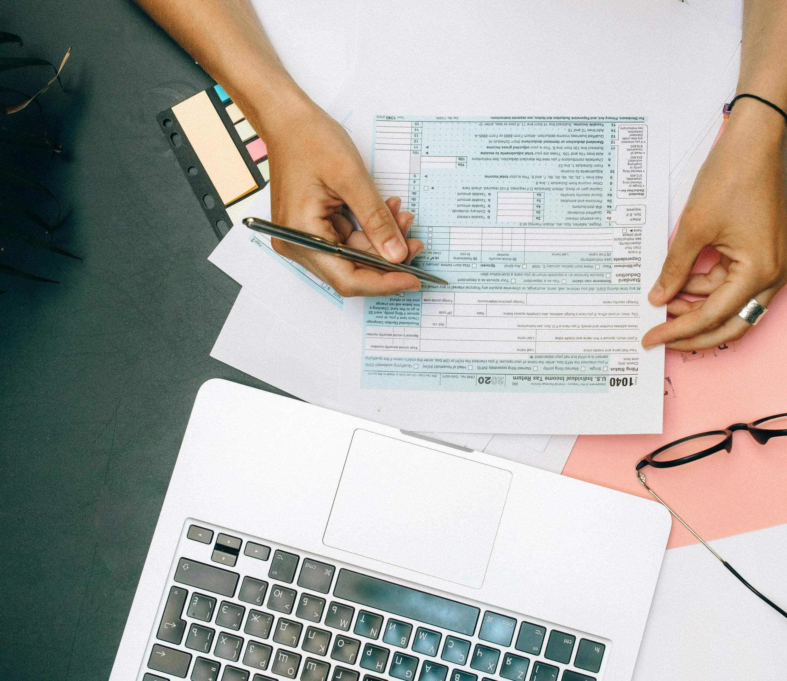 Person filling out tax forms on a desk with a laptop, eyeglasses, a pen, and a color swatch.