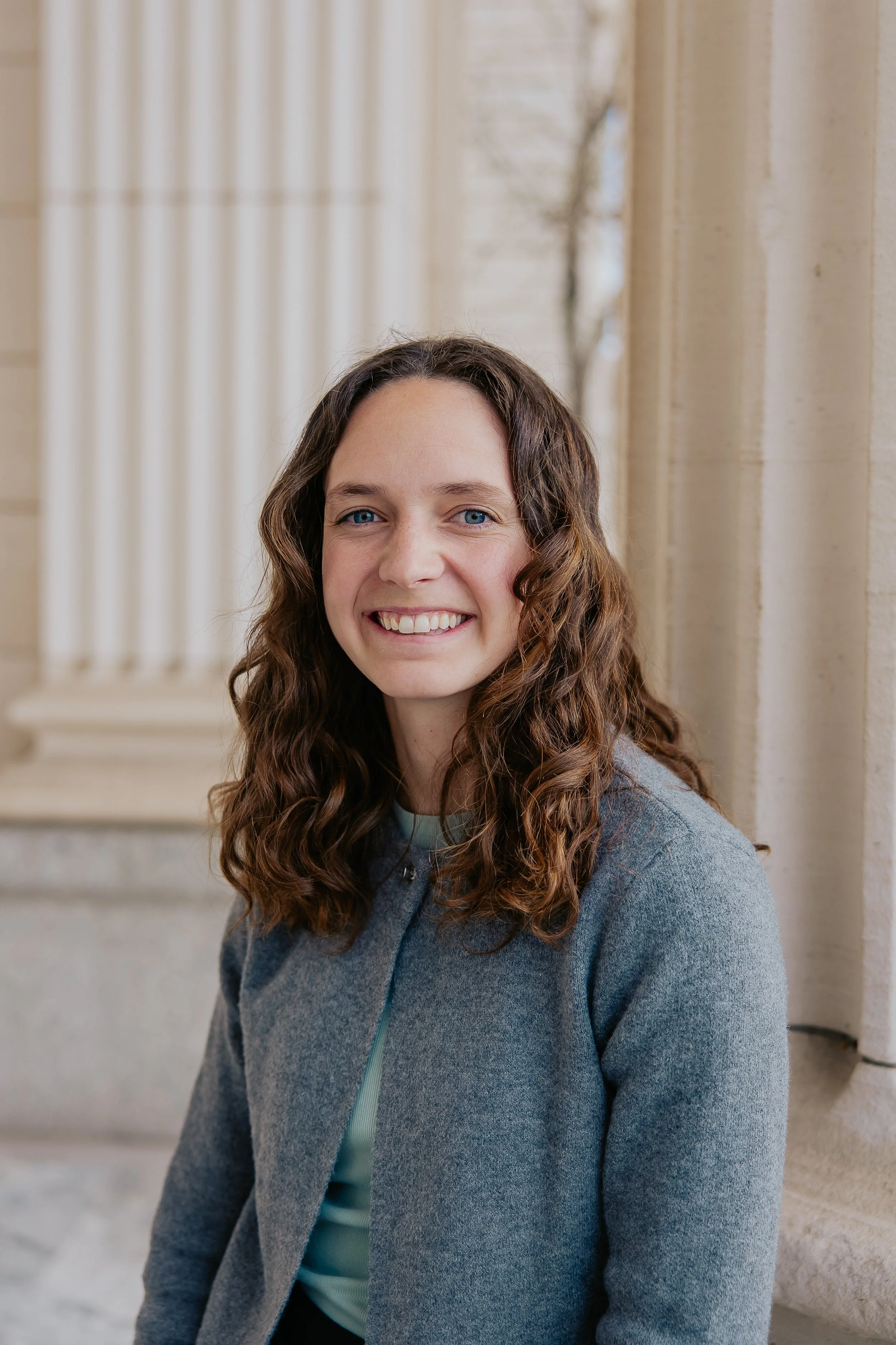 A woman with long, curly brown hair and blue eyes smiling outside a building with stone columns.