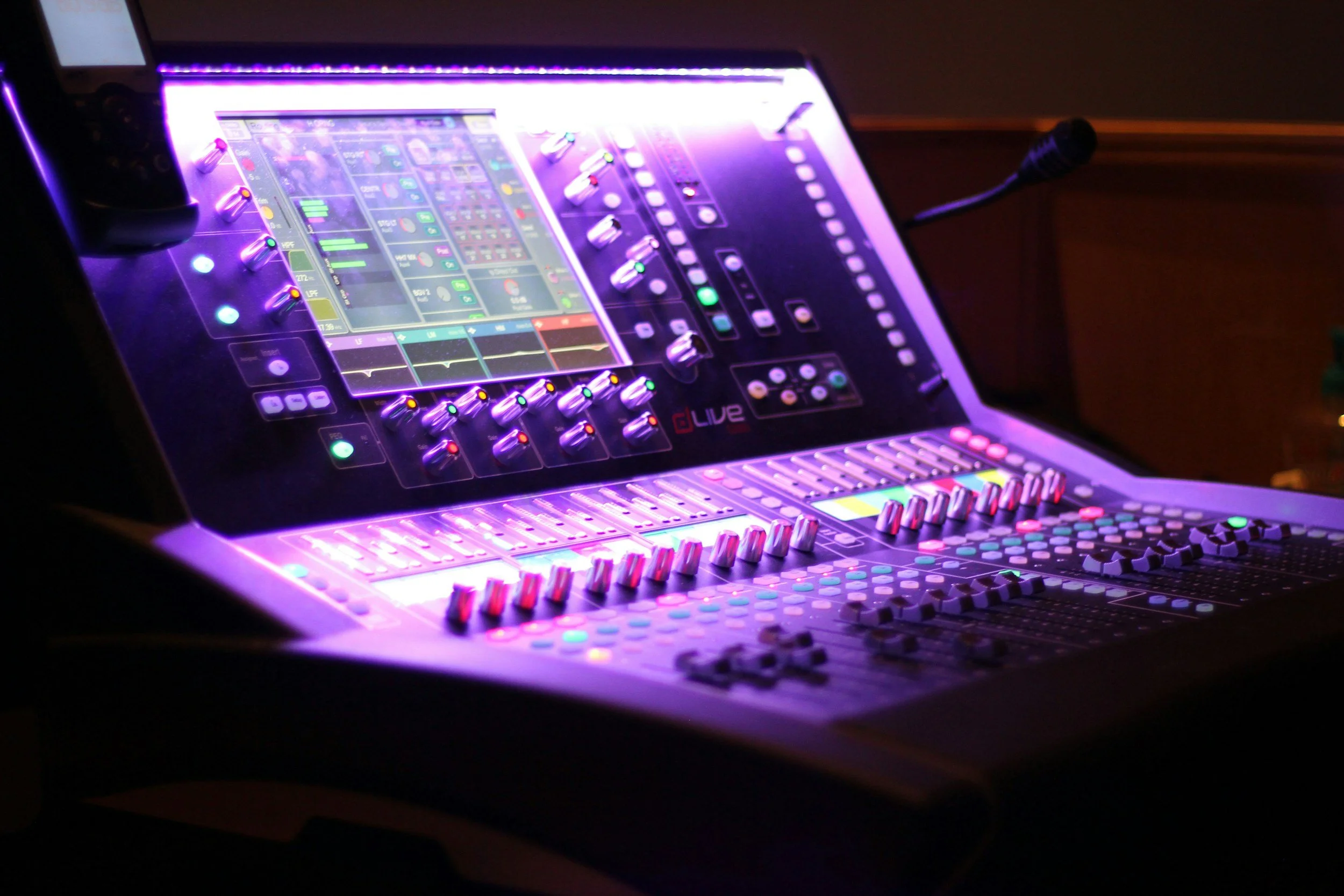 A digital music mixing console with colorful illuminated controls and a screen displaying sound levels, in a dark setting.