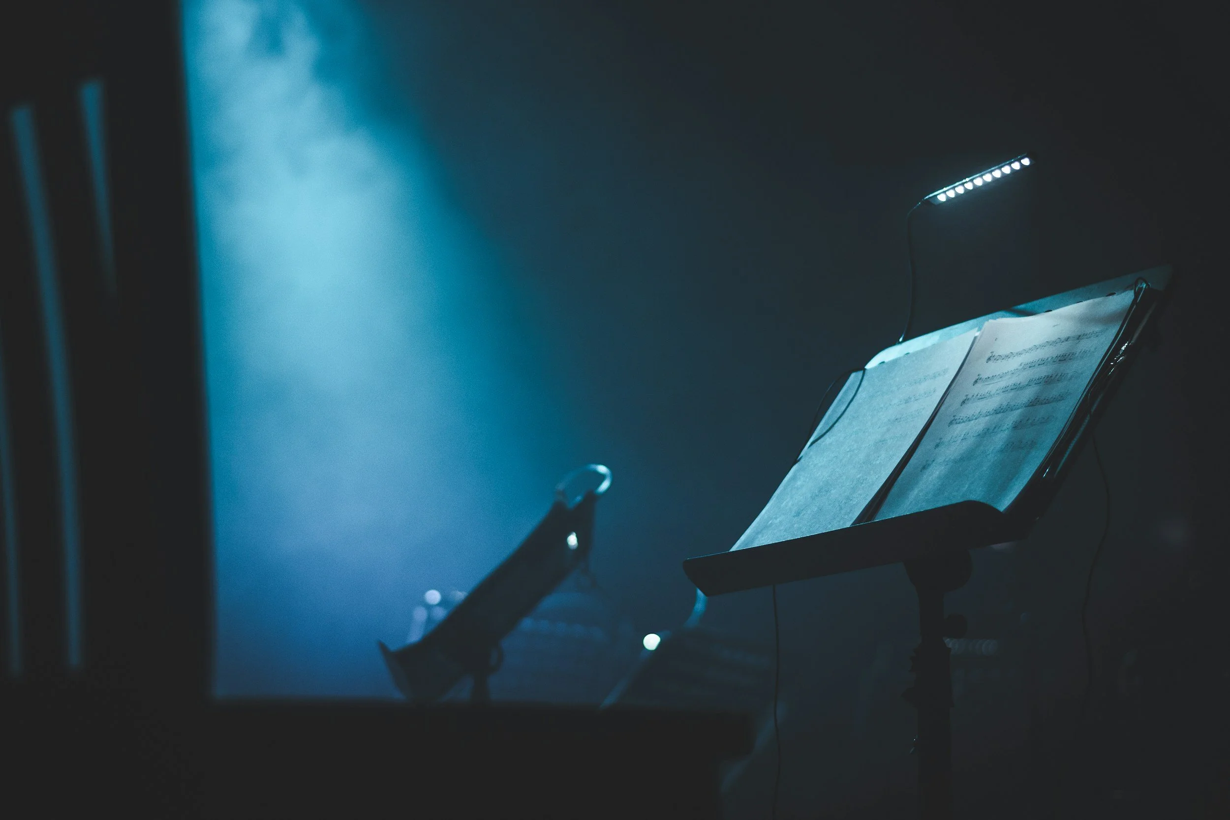 A dimly lit music stand with sheet music illuminated by a small LED light, set against a dark background, indicating a stage or rehearsal space.