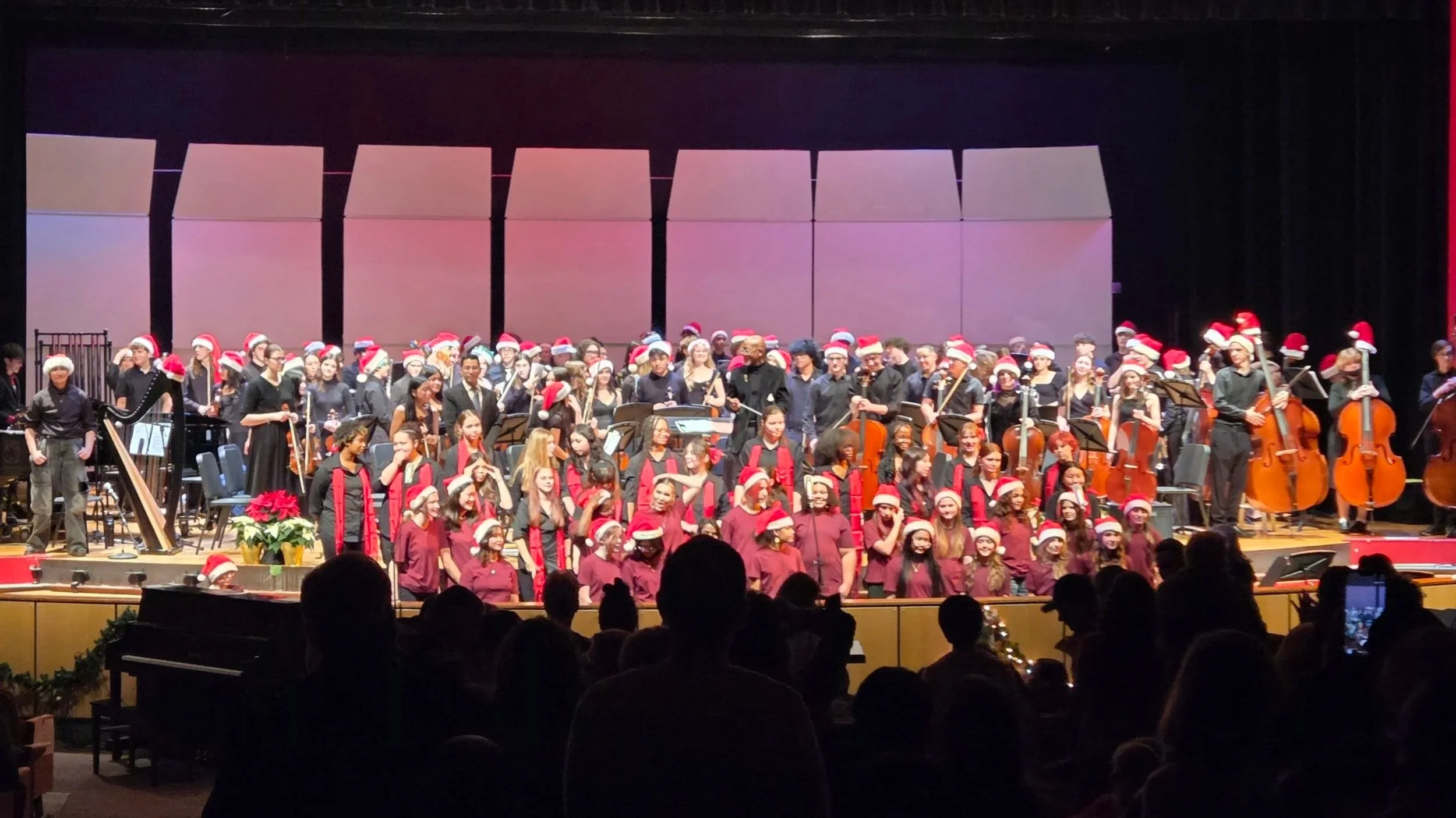 A large choir and orchestra on stage wearing Christmas hats, performing in front of an audience during a holiday concert.