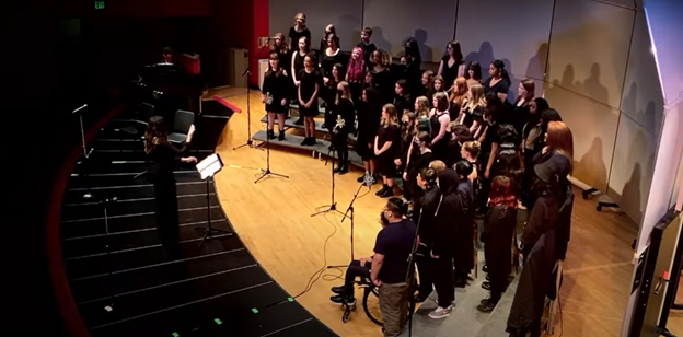 Choir group practicing or performing on stage with a conductor, in a theater or auditorium setting.