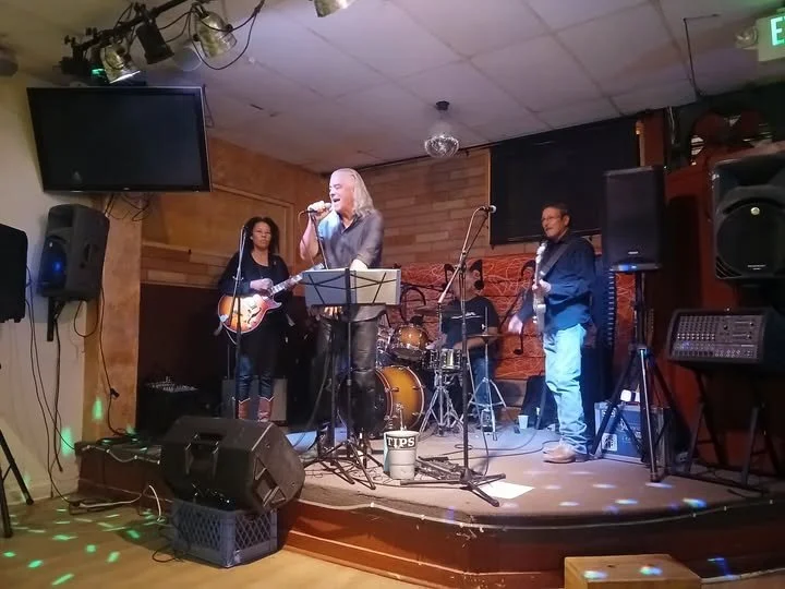 Musicians performing live on stage with a lead singer, guitarist, bassist, and drummer in a cozy venue with wooden walls and stage lighting.